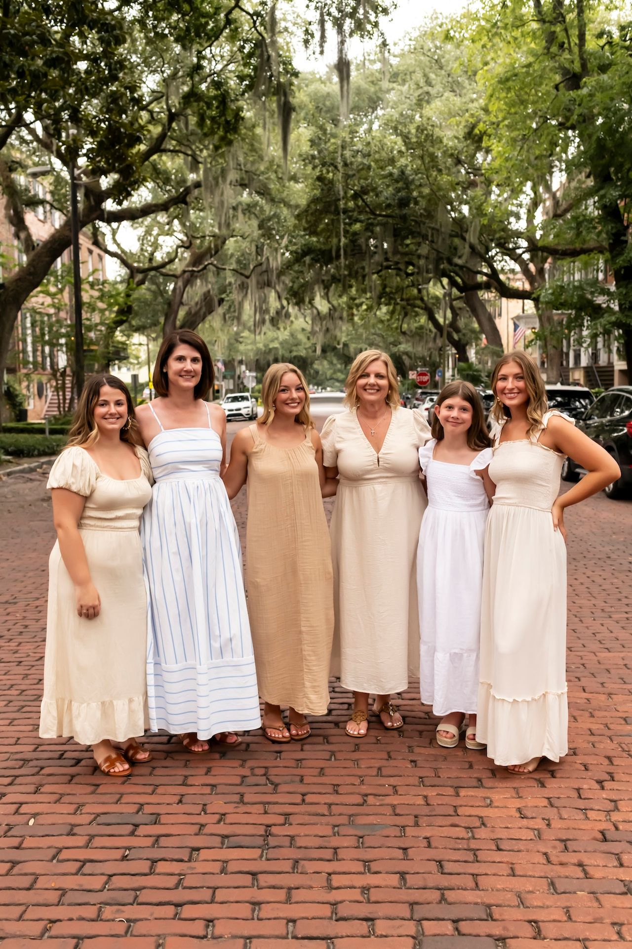 A group of six women wearing light colored maxi dresses stand together on a brick path under trees with Spanish moss.