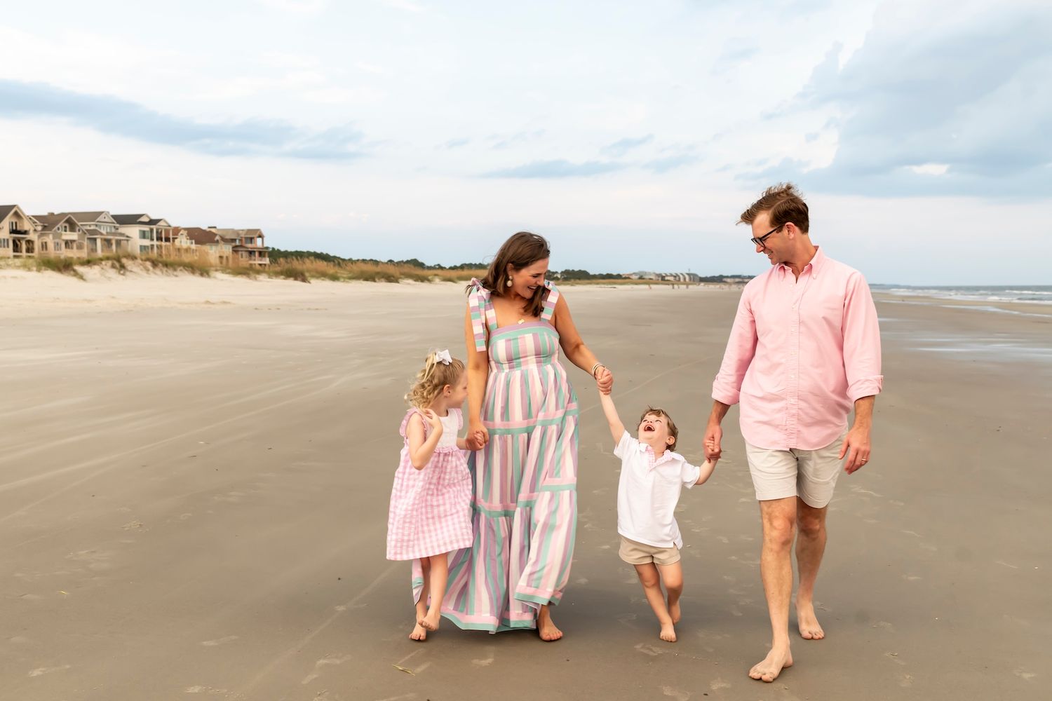 A family in pastel pink and white outfits strolls along a sandy beach at sunset with clouds in the background.