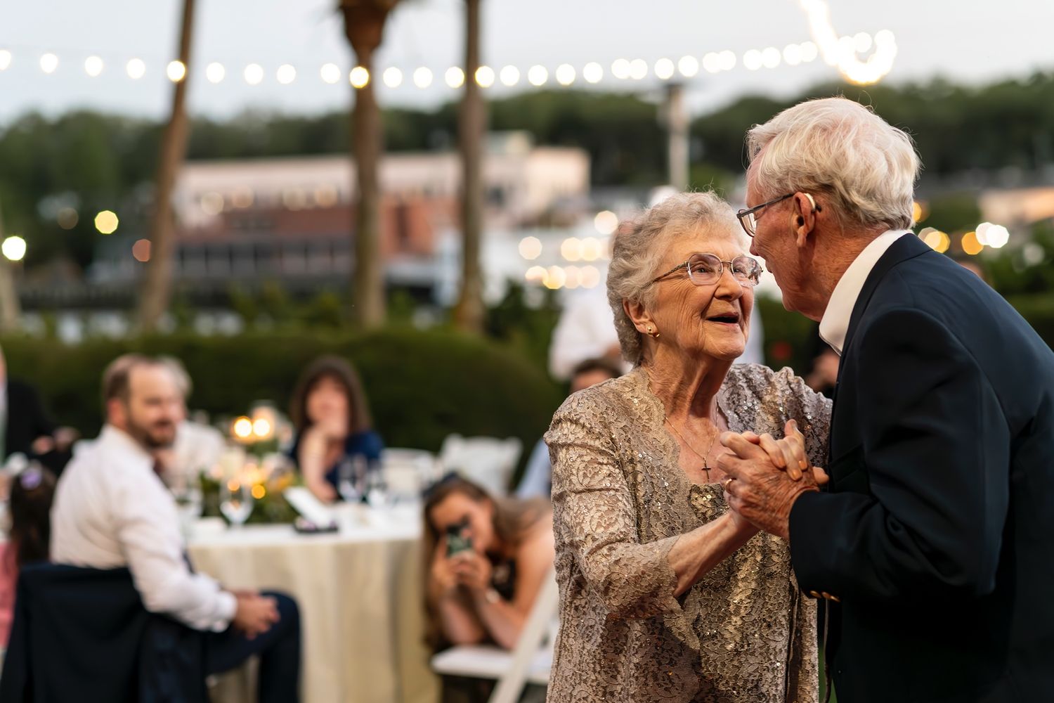 Elderly couple shares a joyful dance together at outdoor evening reception while guests look on from tables.