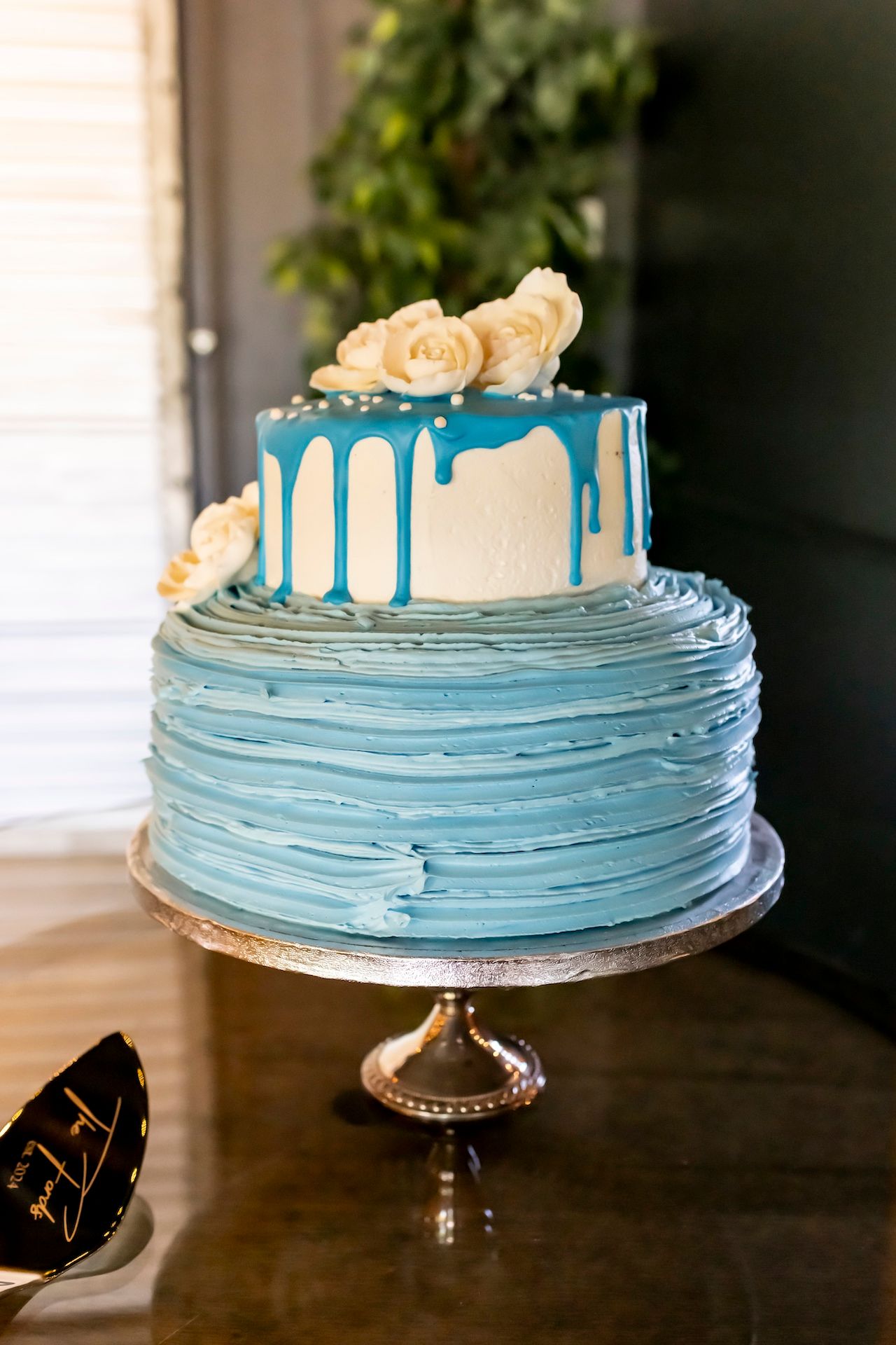 Light blue ombre cake with white chocolate drip and cream roses on silver cake stand features textured buttercream layers.