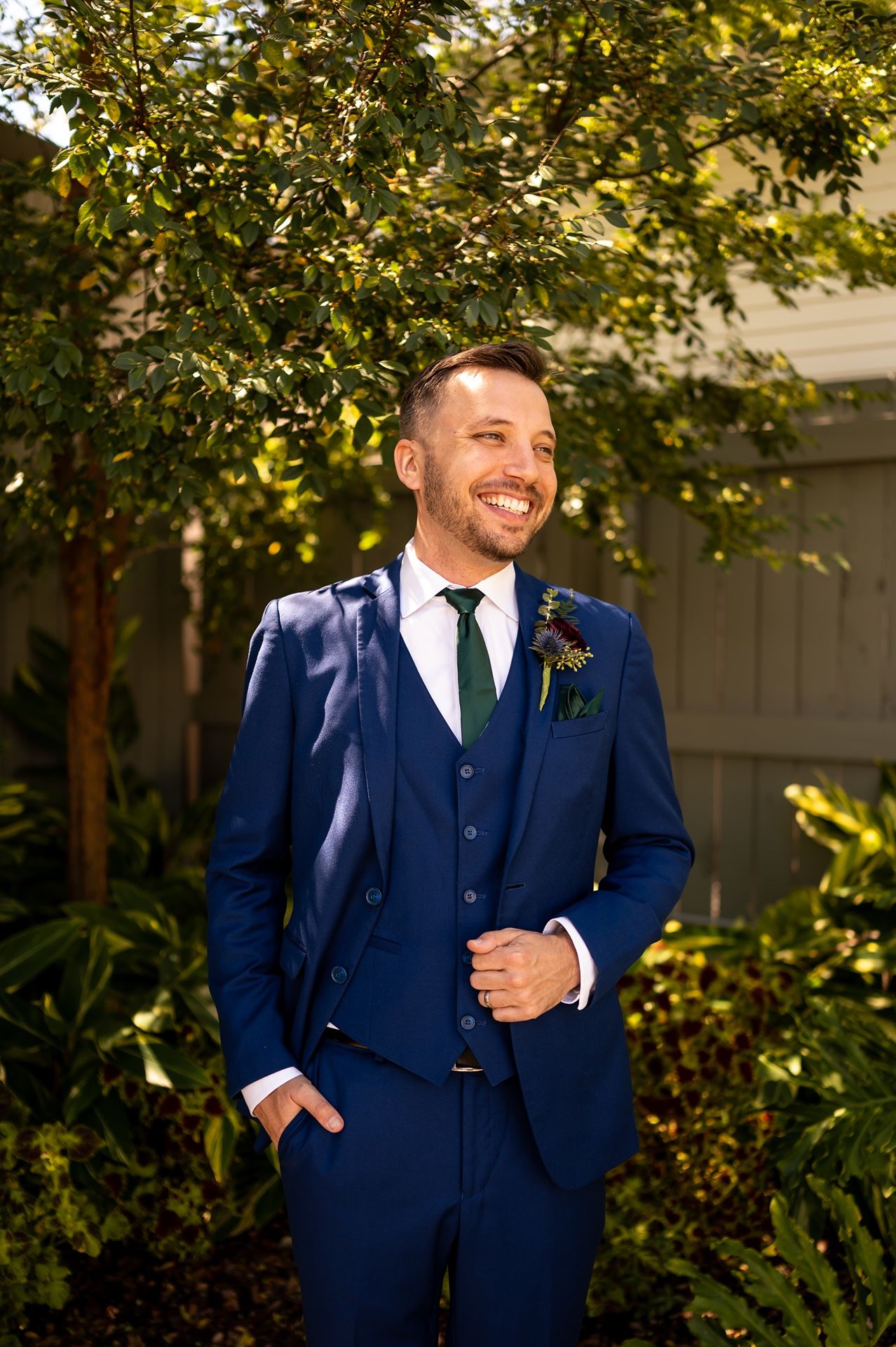 A person in a navy three-piece suit poses outdoors against a backdrop of sunlit trees and foliage.