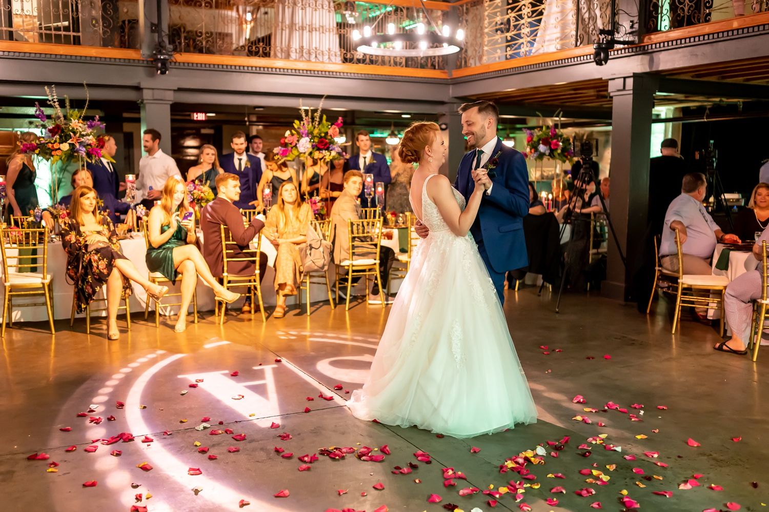 Couple dancing at an elegant wedding reception with rose petals scattered on illuminated dance floor and guests watching.