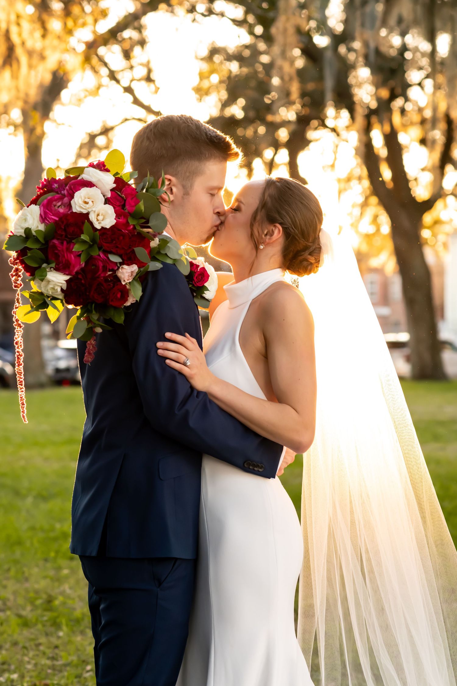 Couple shares a romantic sunset kiss wearing wedding attire in a grassy park with golden light filtering through trees.