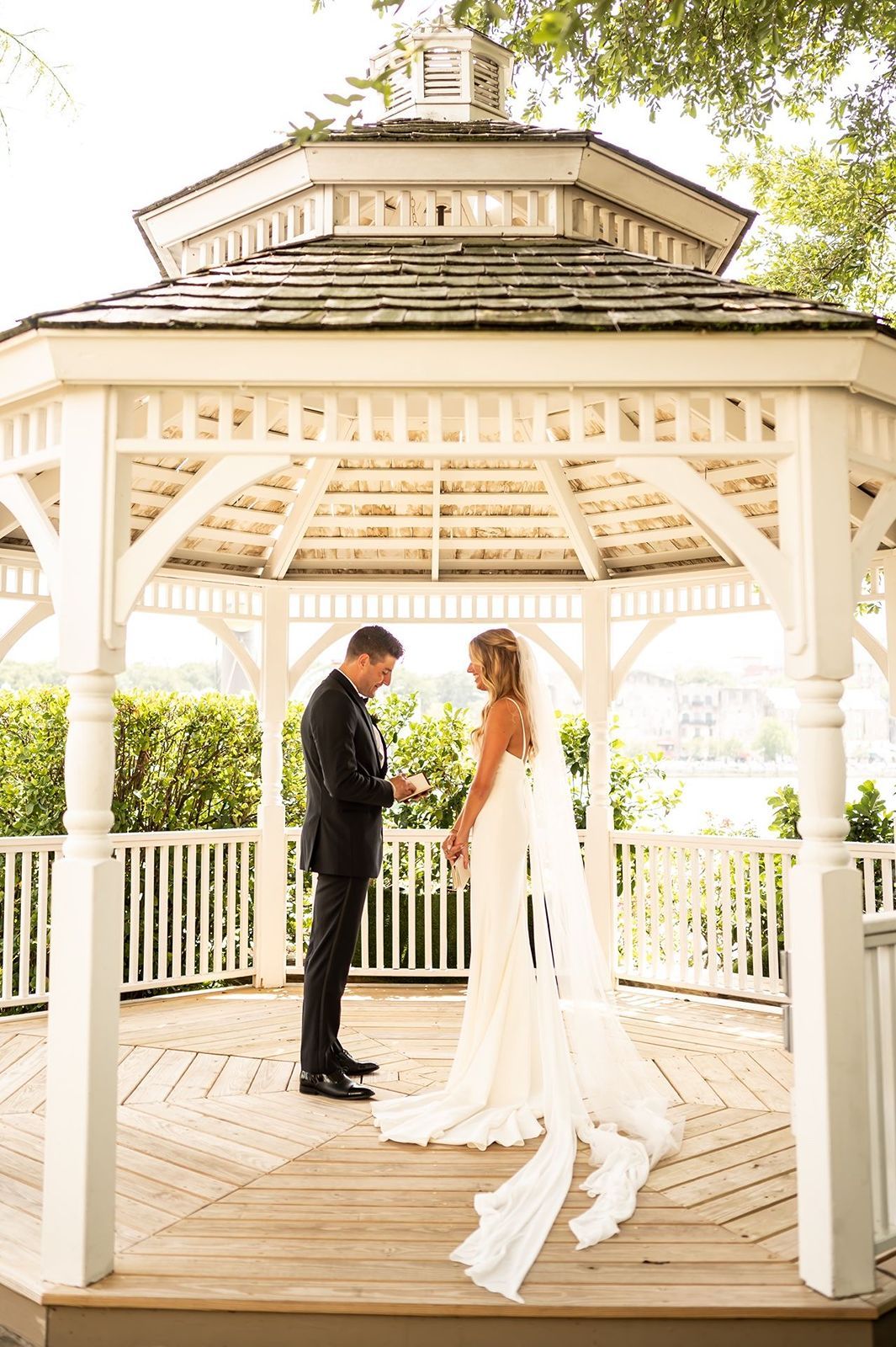 A romantic moment between a couple in elegant attire standing beneath a white wooden gazebo on a sunny afternoon.