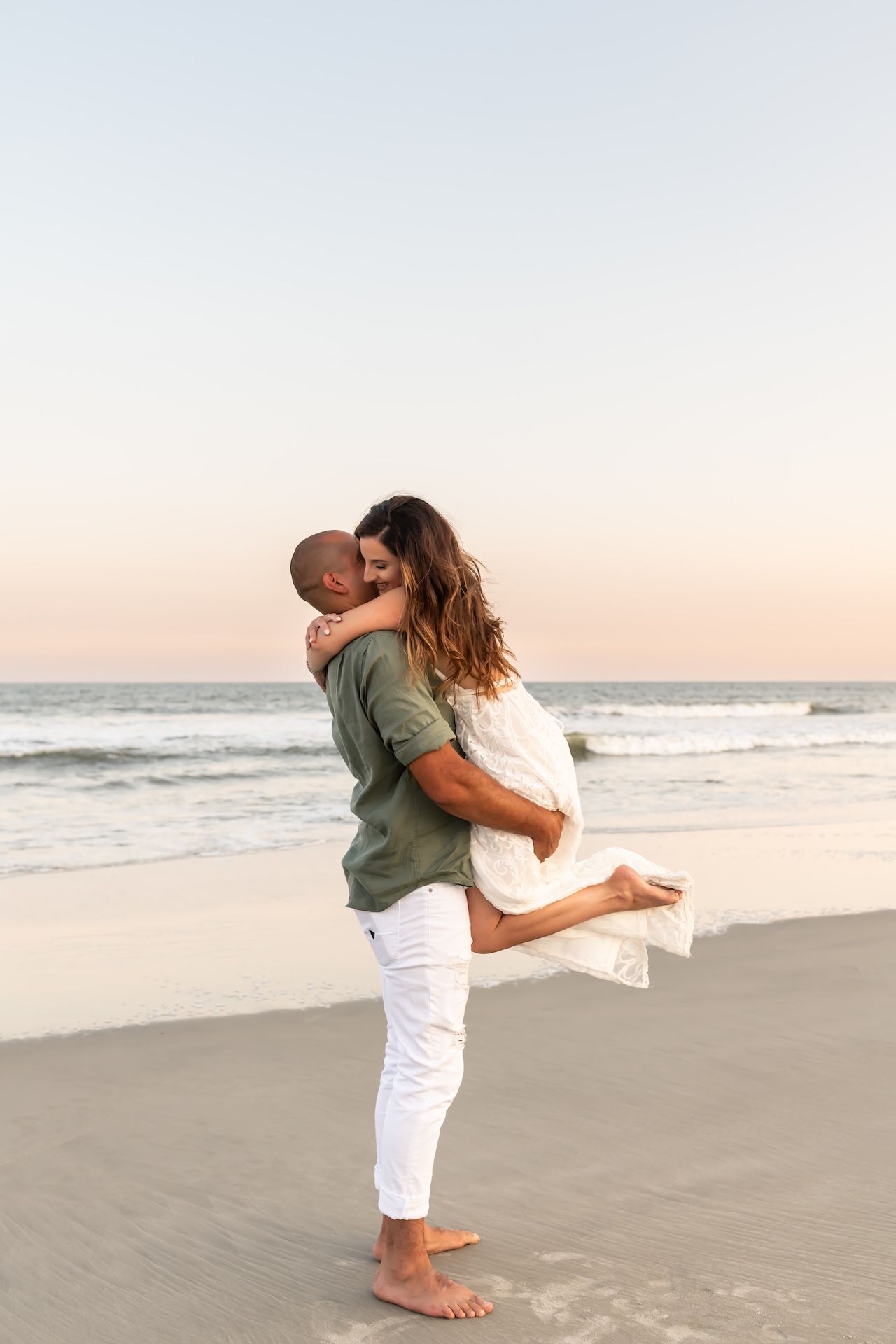 A romantic couple embraces on a sandy beach at sunset with ocean waves in the background.