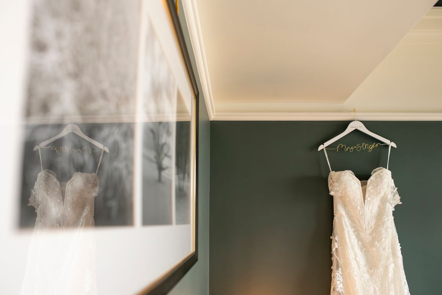 White wedding dress hanging on a hanger against a dark green wall in natural lighting.