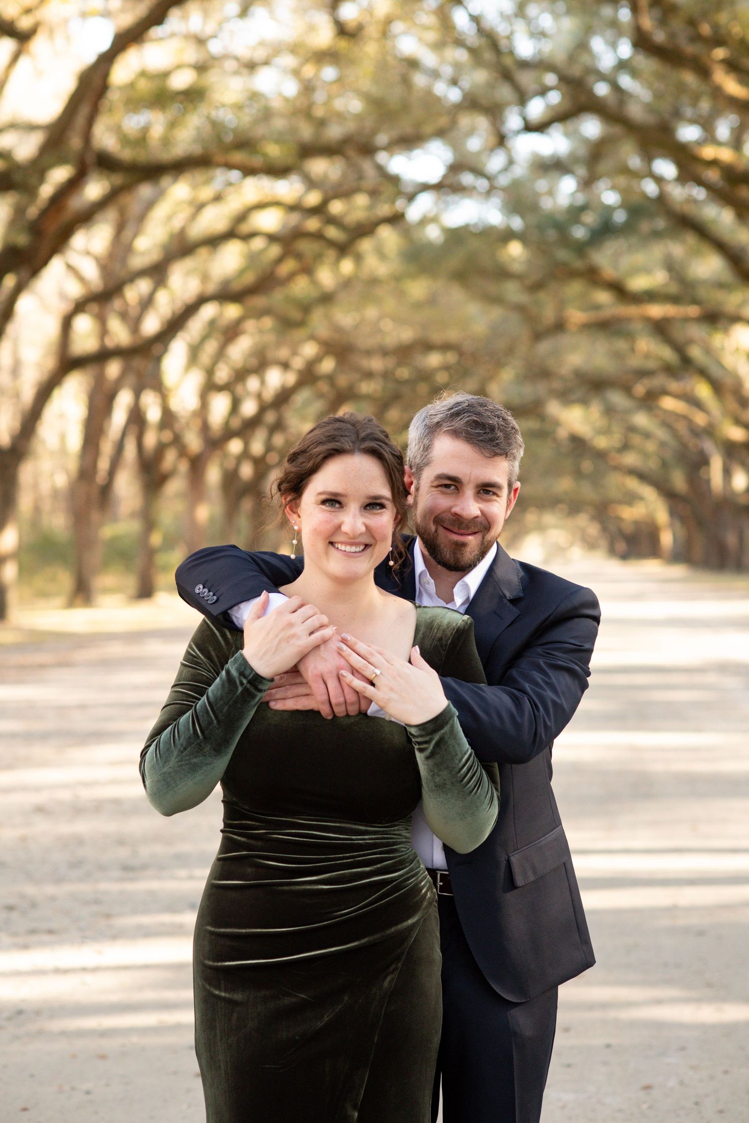 Couple embracing on a tree-lined path, woman in green velvet dress, man in navy suit, smiling outdoors.