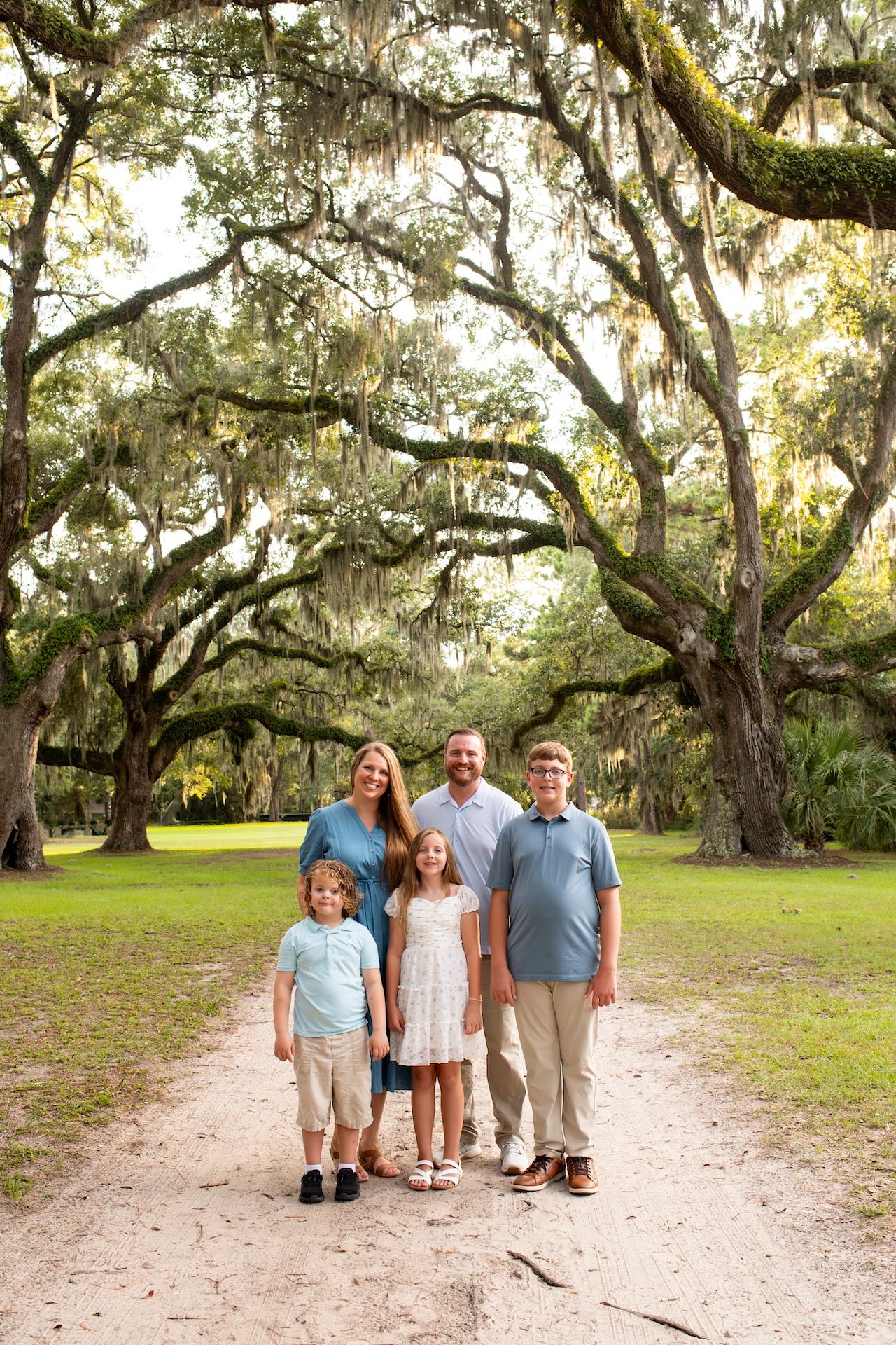 Family of five posing on a sandy path under sprawling moss-draped oak trees in a lush green Southern landscape.