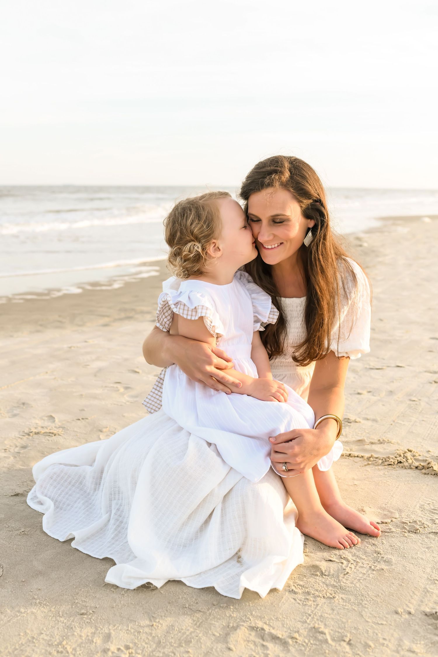 Tender moment between mom and daughter in white dresses sitting on beach at sunset.