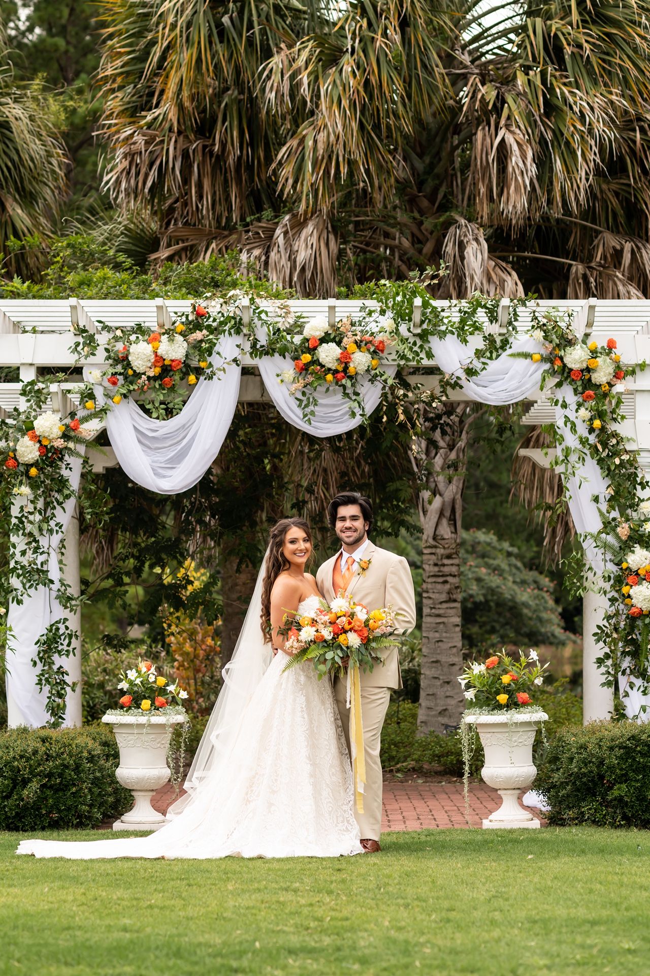 Wedding ceremony setup with white pergola arch decorated with lush flowers and white draping under palm trees.