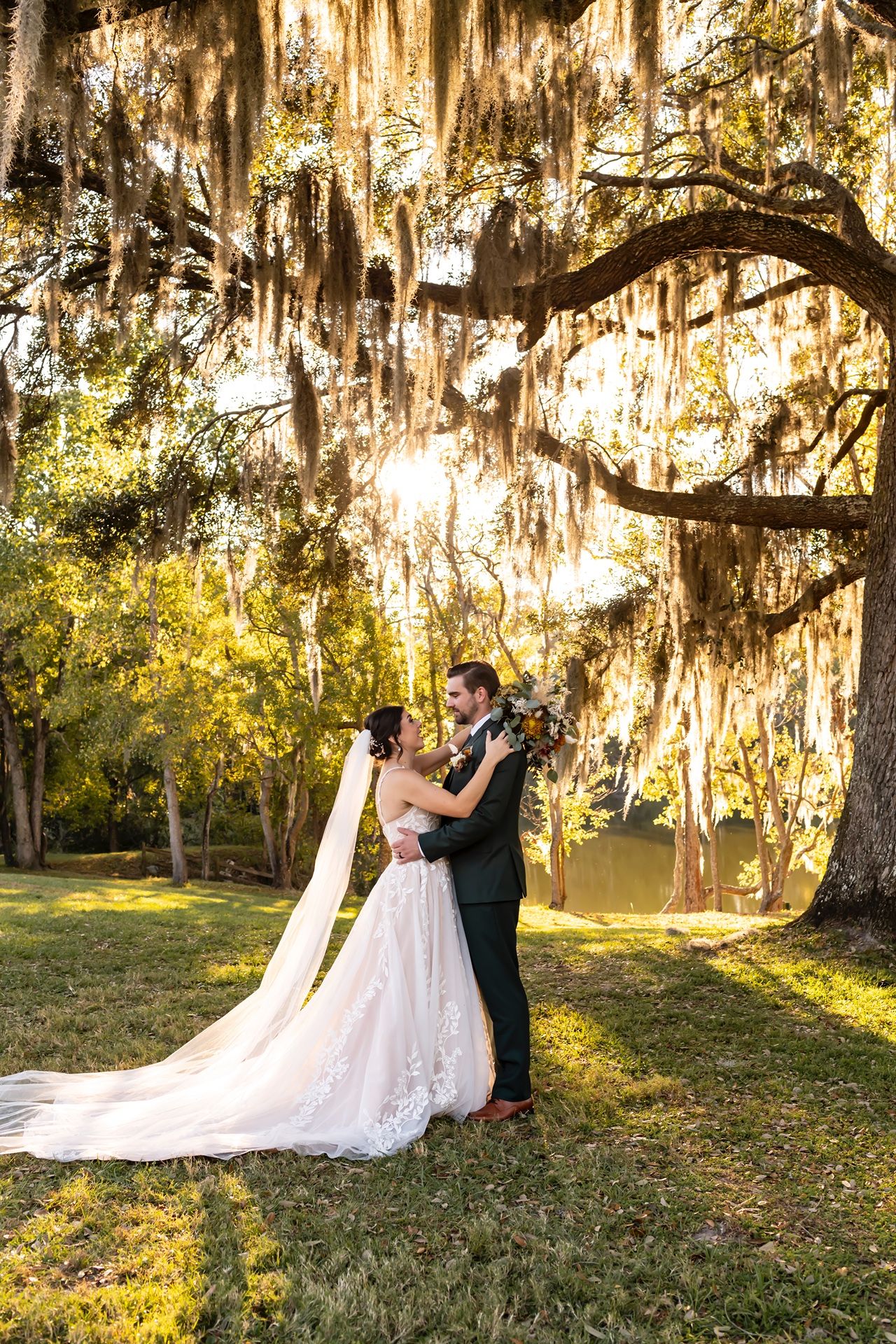 A couple shares a romantic embrace under Spanish moss draped trees at sunset in a dreamy outdoor setting.