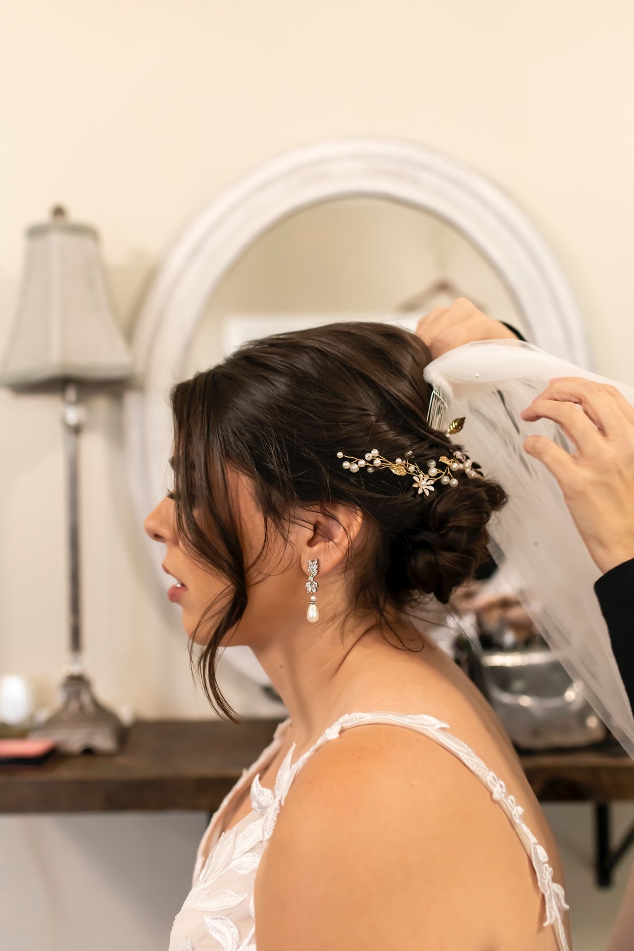 A bride having her veil and headpiece adjusted in a getting ready room with natural lighting.