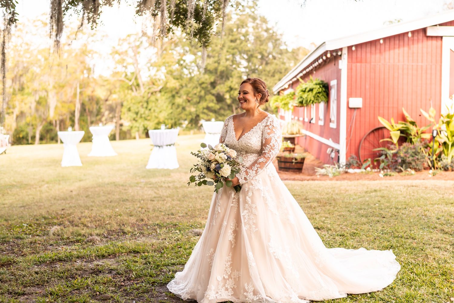 Bride wearing lace gown with flowing skirt poses with bouquet outside red barn on sunny autumn afternoon.