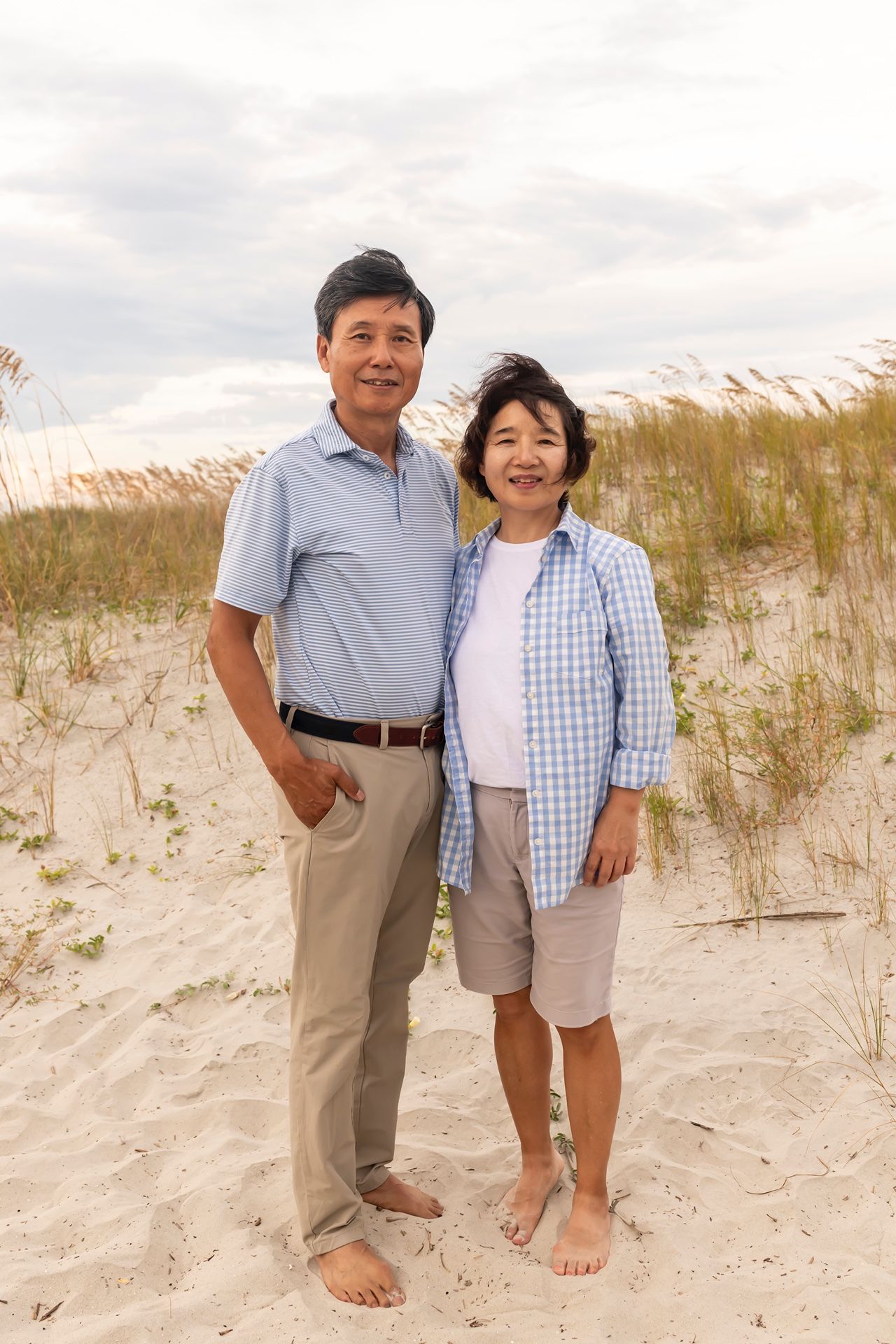 A couple stands together barefoot on sandy dunes with beach grass and cloudy skies behind them.