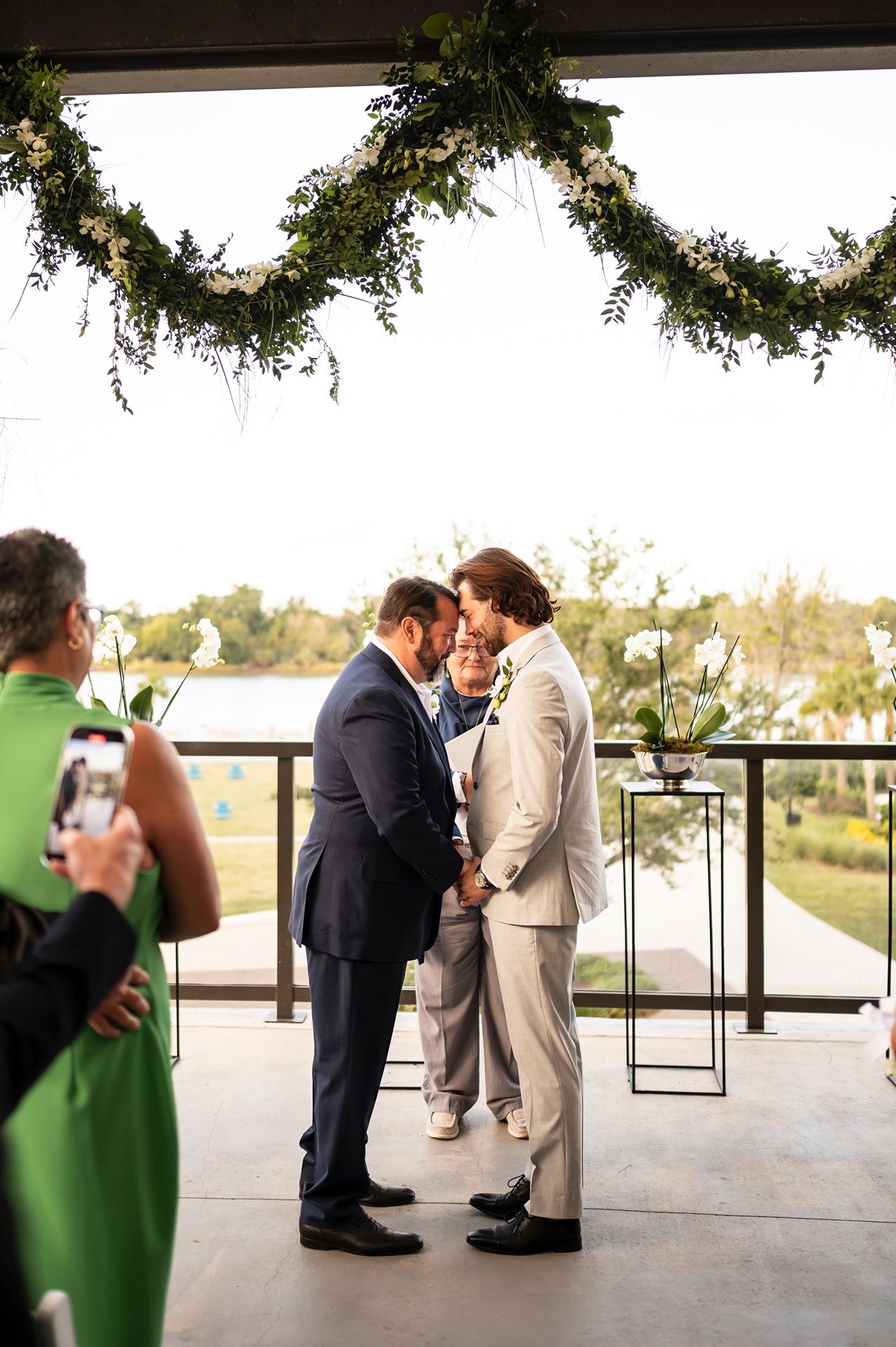 Romantic wedding couple embracing and kissing during outdoor terrace ceremony with floral archway decorations.