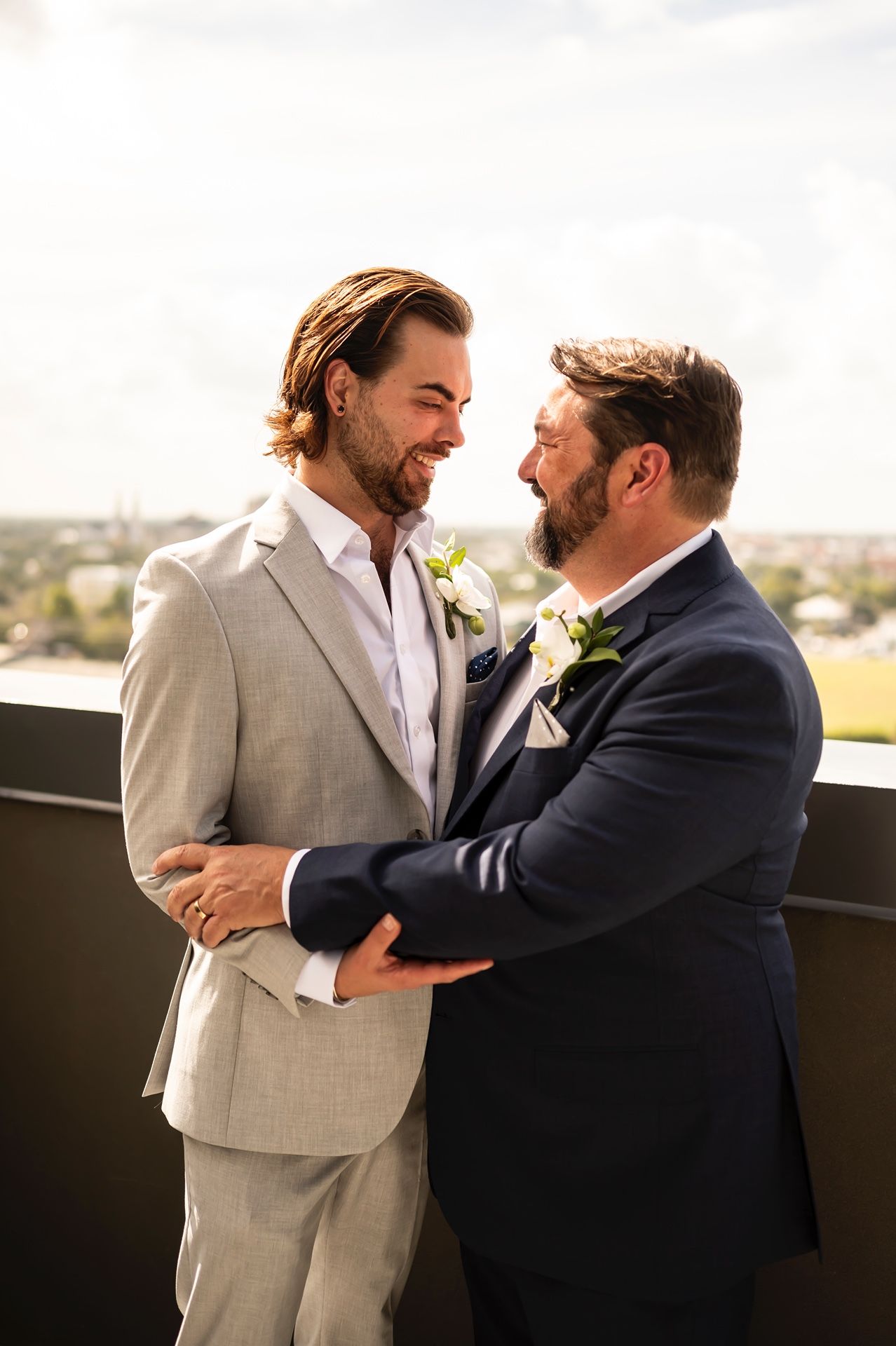 Two people in formal suits share a joyful hug and moment of laughter outdoors on a sunny day.