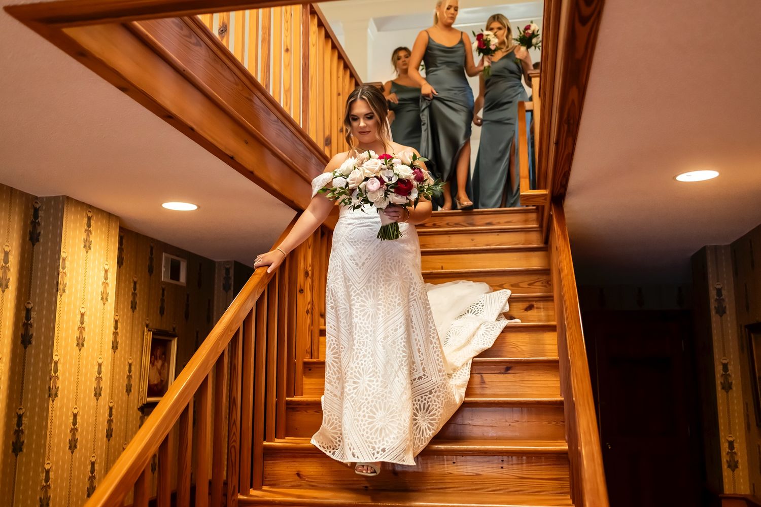 People descending a wooden staircase in formal attire at an indoor venue with someone in a white dress leading the way.