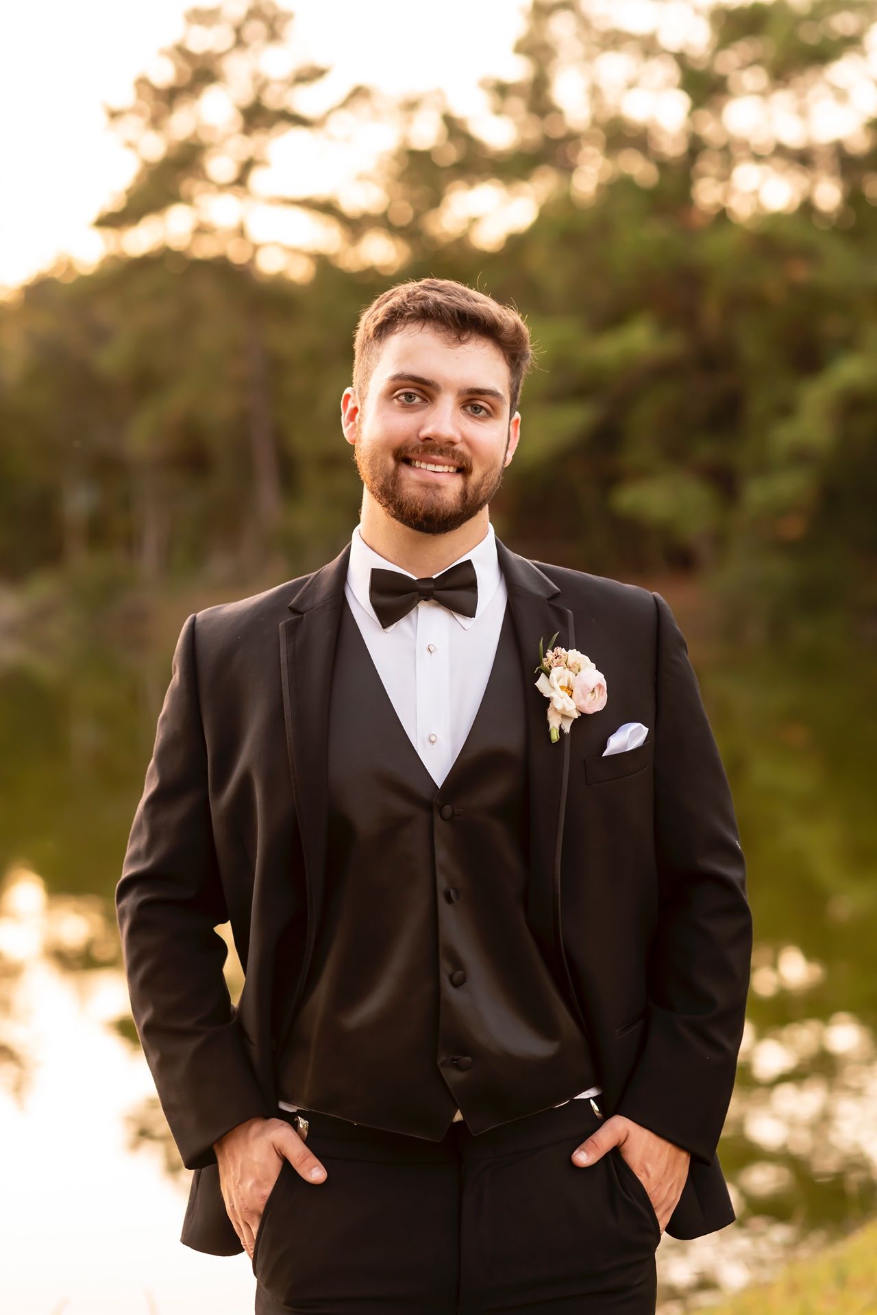Groom in black three-piece tuxedo with bow tie and boutonniere stands outdoors at sunset with trees in background.