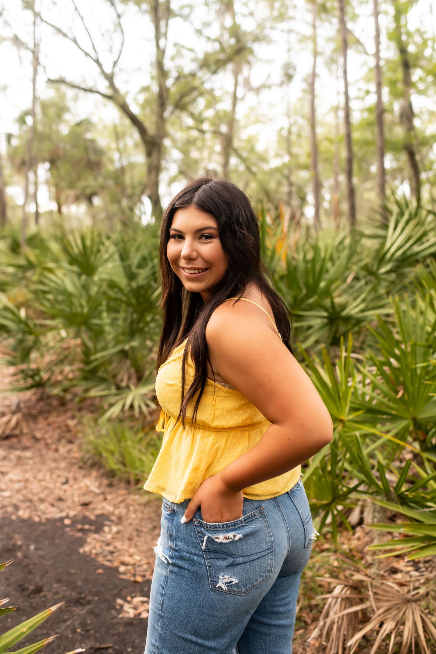 Woman in yellow sleeveless top and ripped jeans poses in front of palm plants in natural outdoor setting.