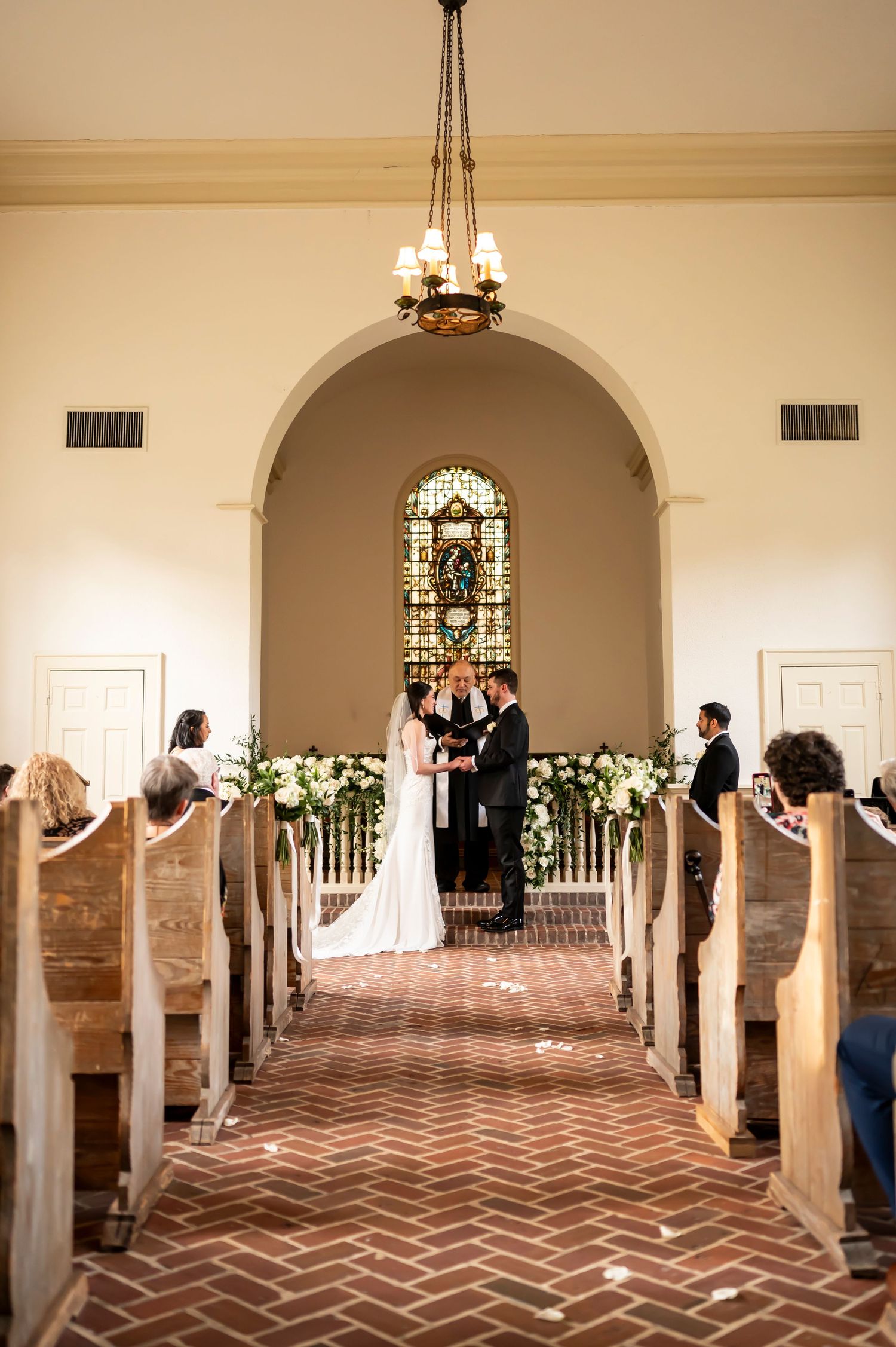 Traditional church wedding ceremony inside a chapel with a stained glass window and herringbone brick flooring.