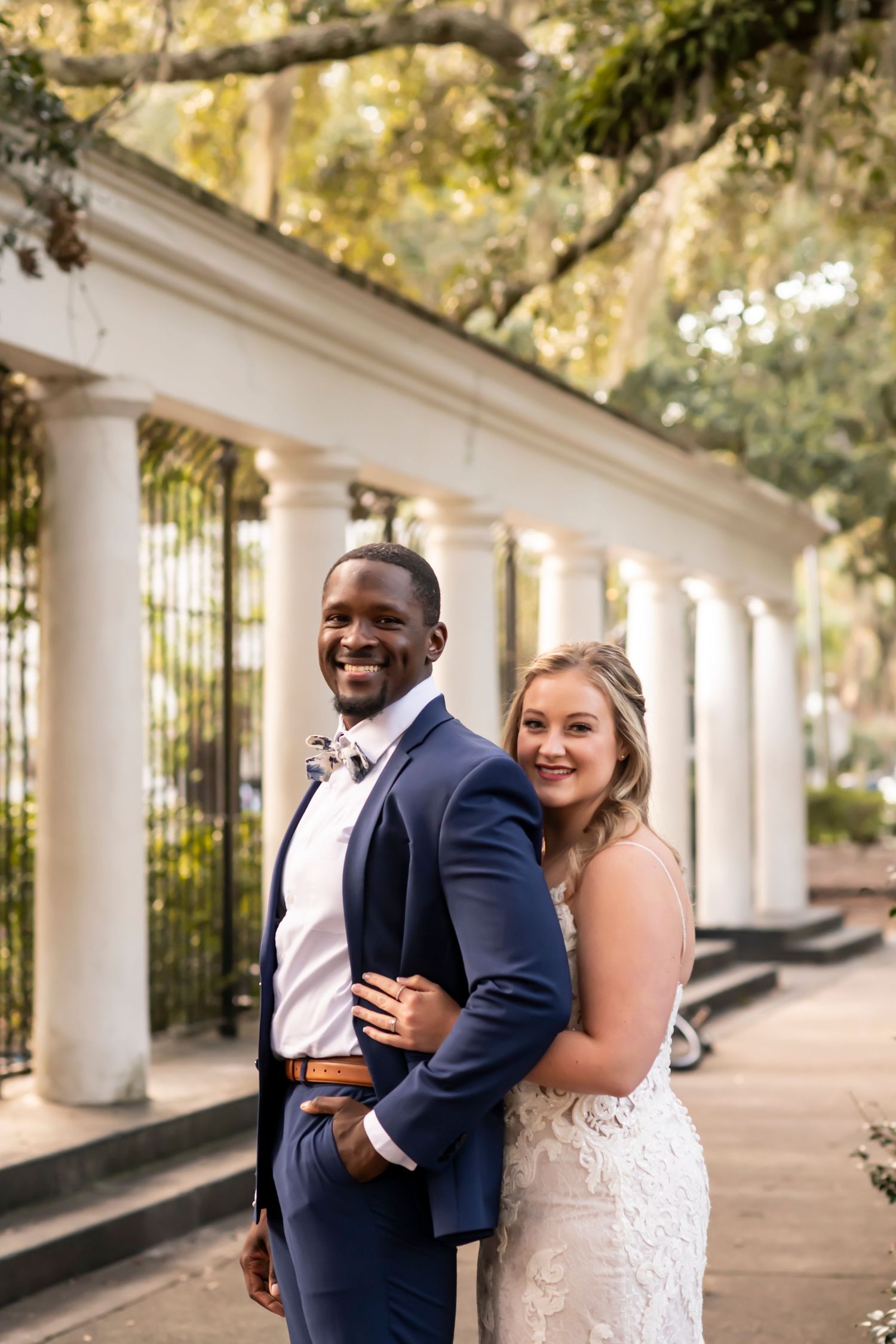 A couple in formal wedding attire poses together by white columns and Spanish moss on their special day.