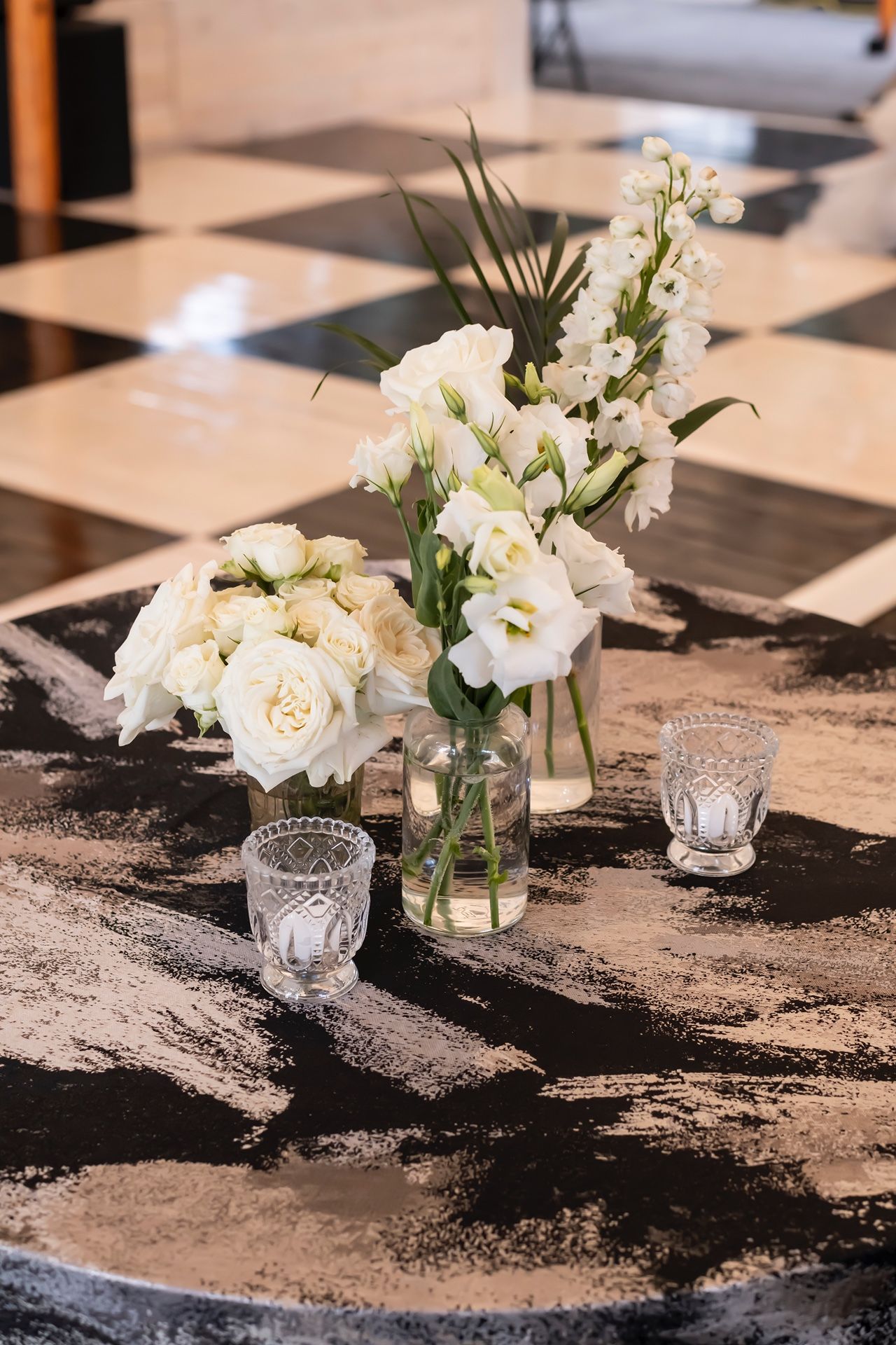 White floral centerpiece with roses and delicate blooms in a glass vase on a marble-effect table with crystal votives.
