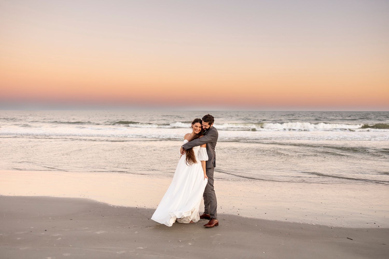 A couple in formal attire embraces on a sandy beach at sunset with ocean waves in the background.