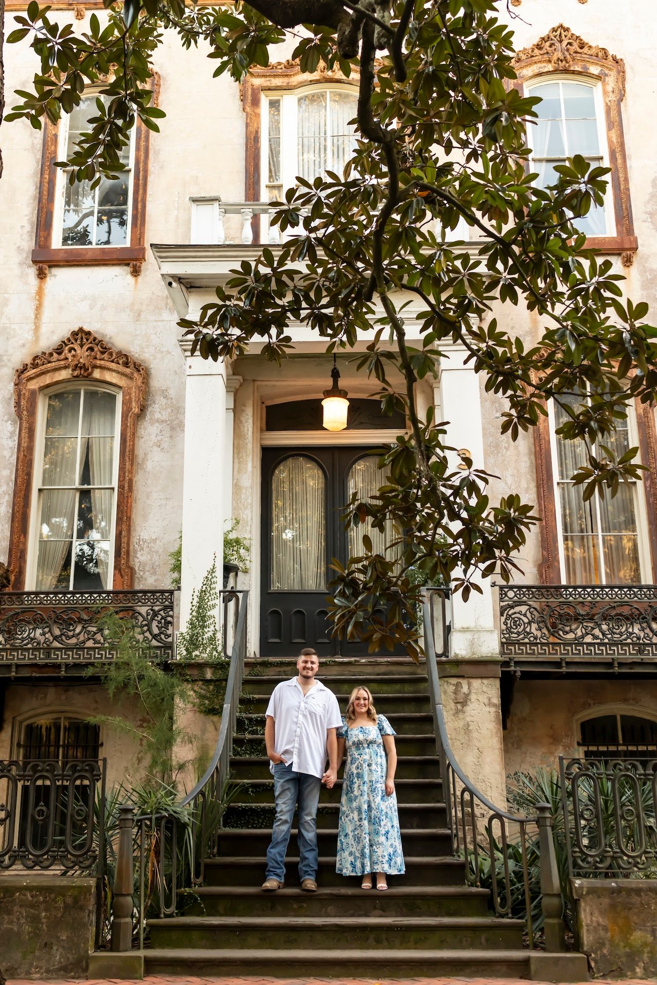 Two people stand on the steps of a historic Southern mansion with ornate decorative elements and magnolia trees.