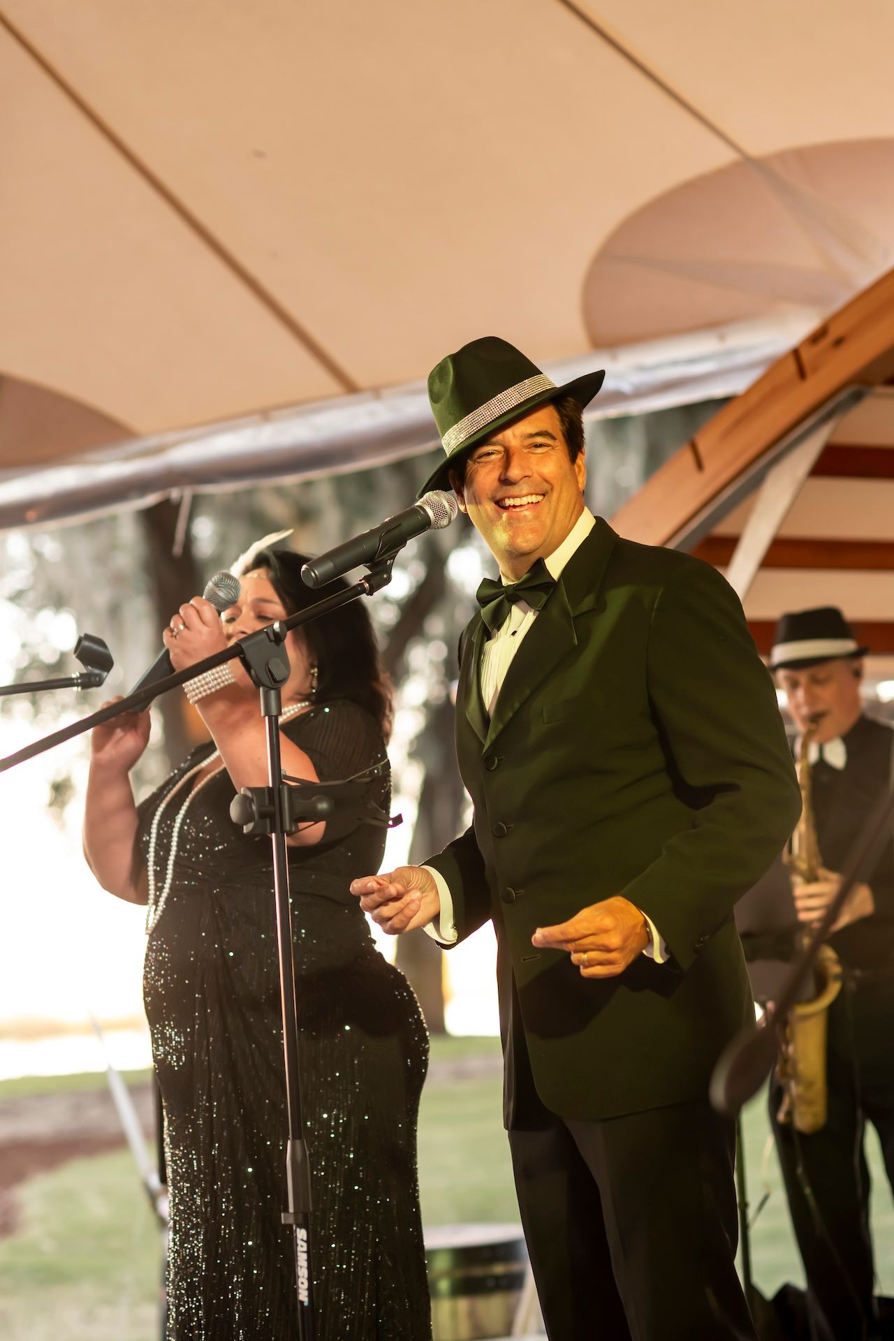 A performer in a black tuxedo and fedora hat smiles while singing at an elegant outdoor event with a band.