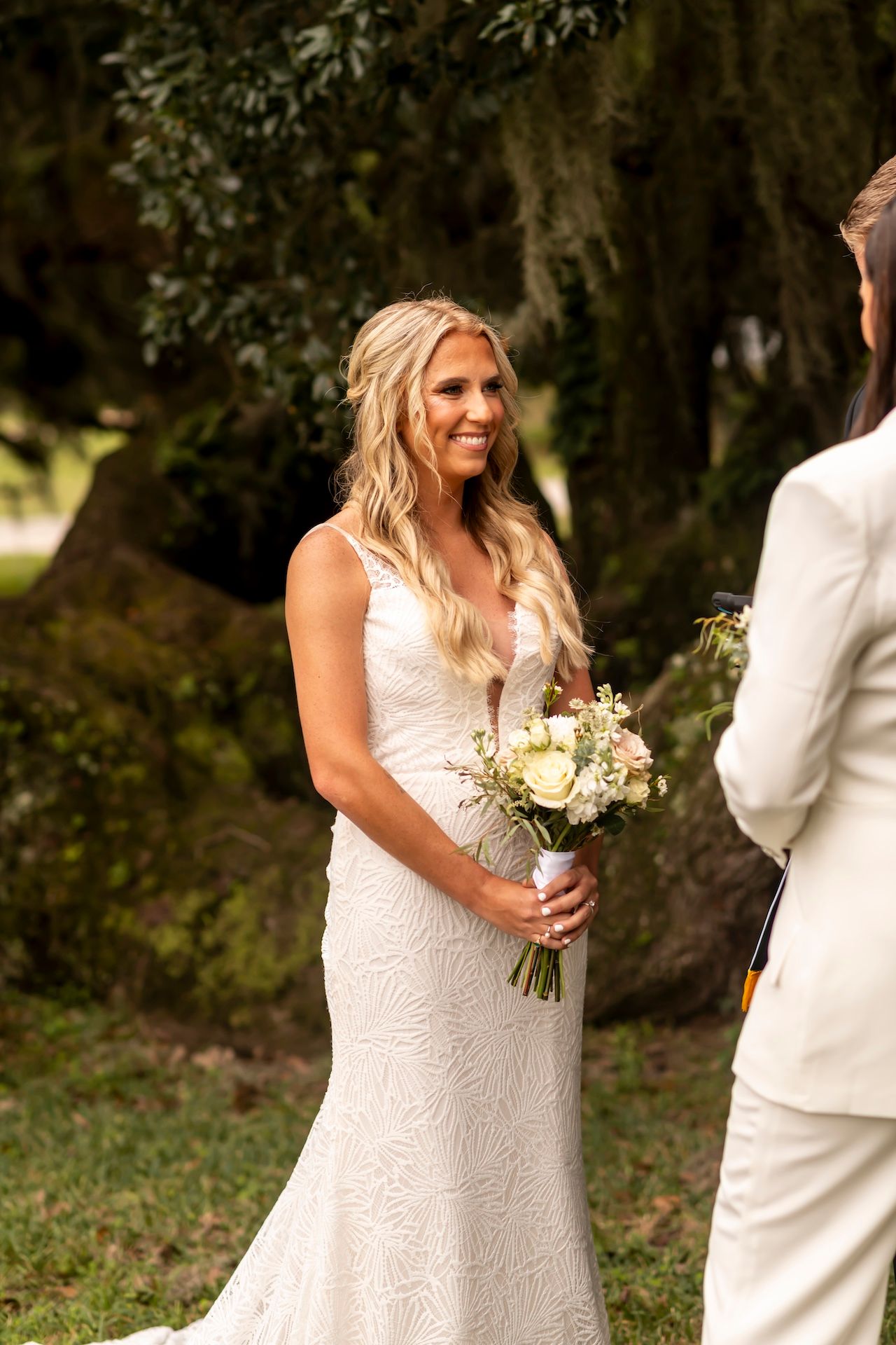 Bride in lace wedding dress holding white bouquet smiles during outdoor ceremony with moss-draped trees.