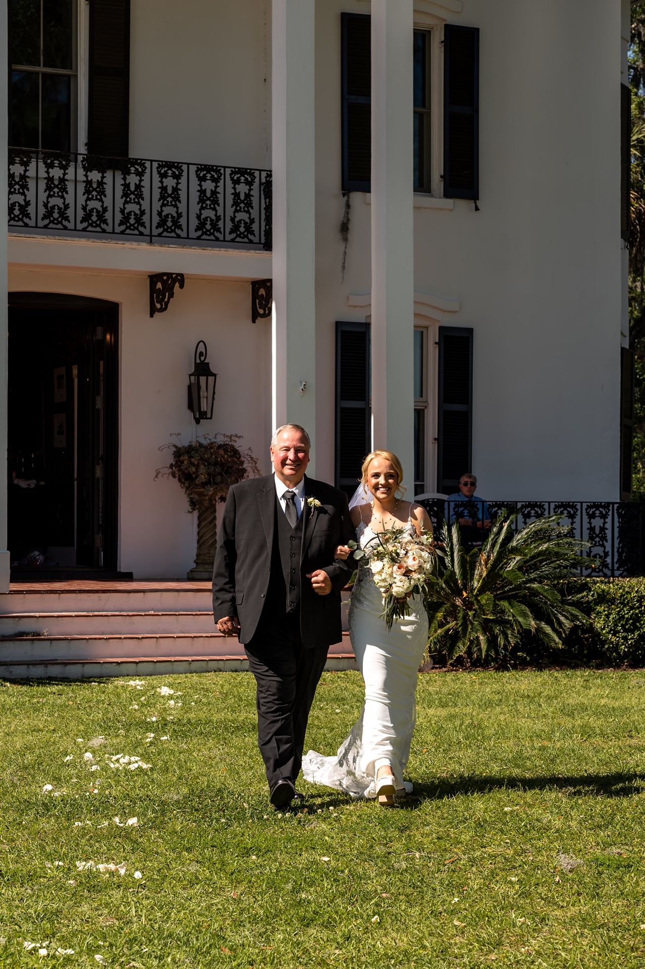 A wedding couple walks on a lush lawn in front of a grand Southern mansion with white columns and ornate iron railings.