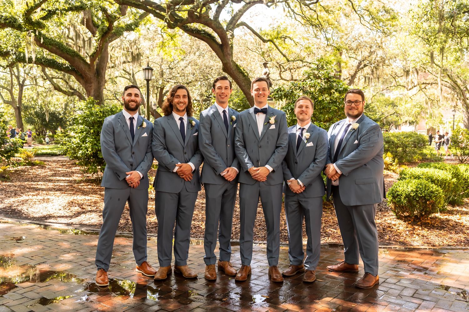 Groomsmen in charcoal gray suits laughing together in an outdoor garden setting during a wedding photoshoot.