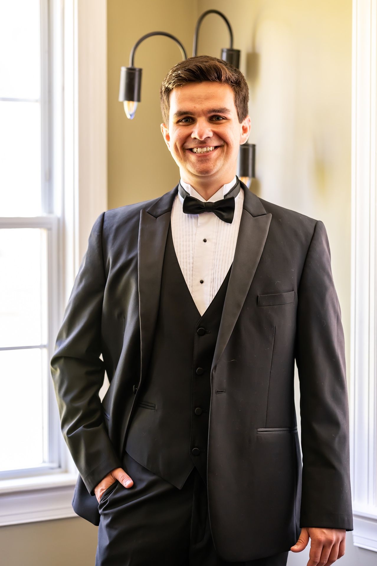 Person in formal black tuxedo with bow tie smiles while posing in bright indoor setting.