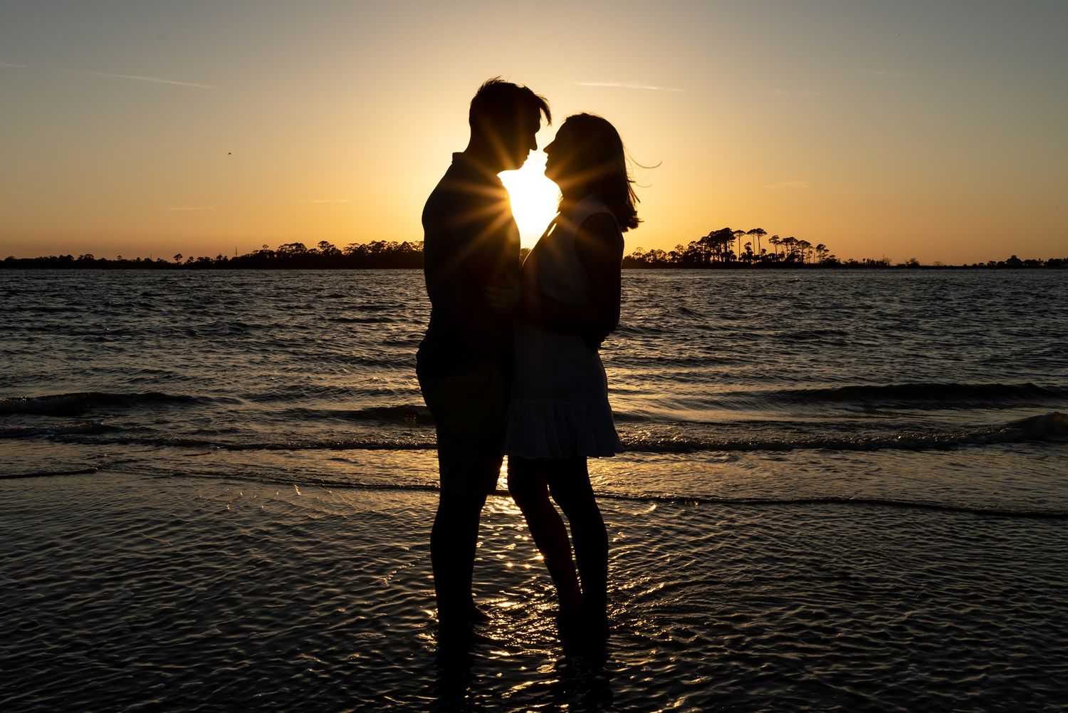 A silhouetted couple embraces on a beach at sunset with golden light streaming through their figures.