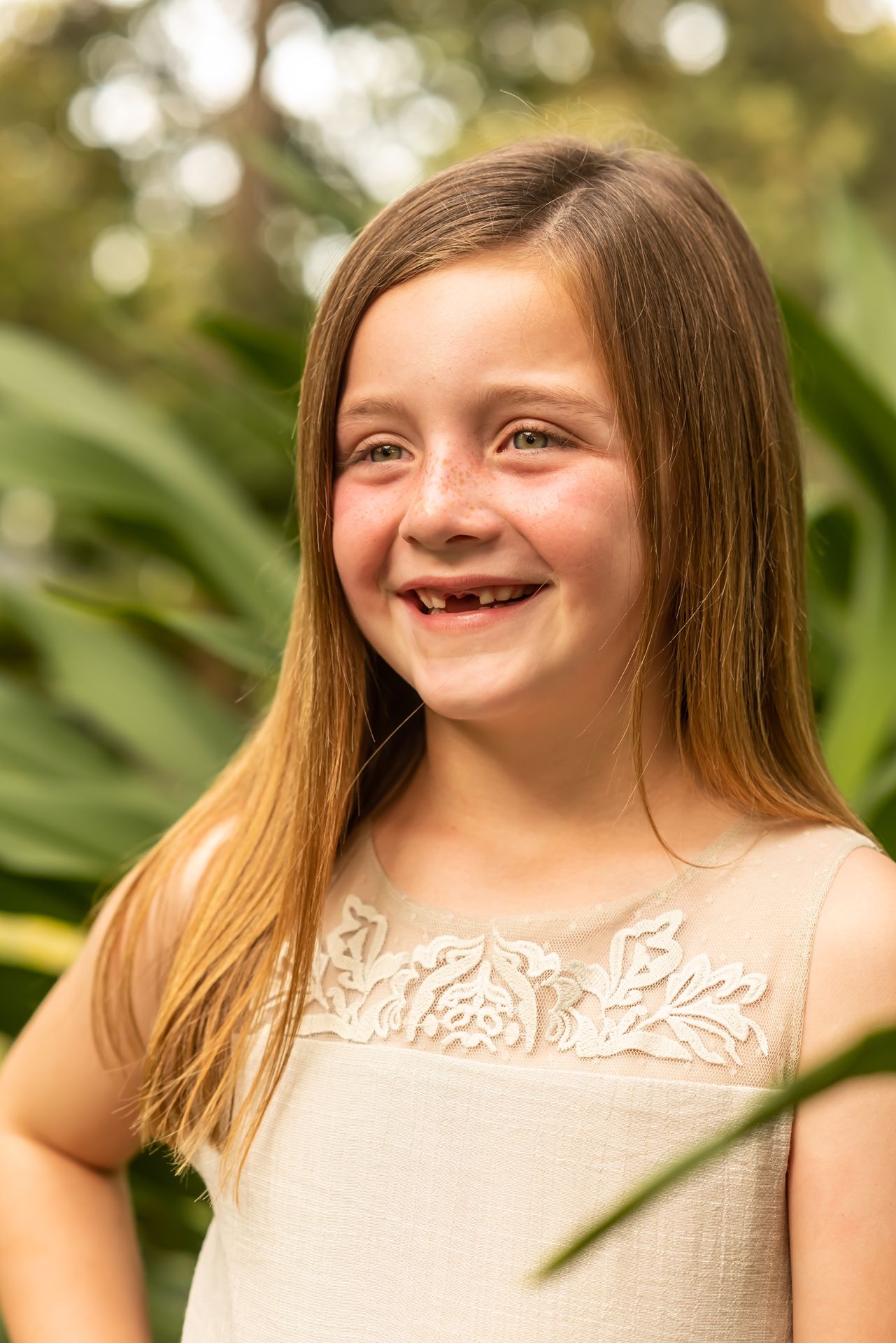 A child wearing a light colored lace dress smiles joyfully against a soft green outdoor background.