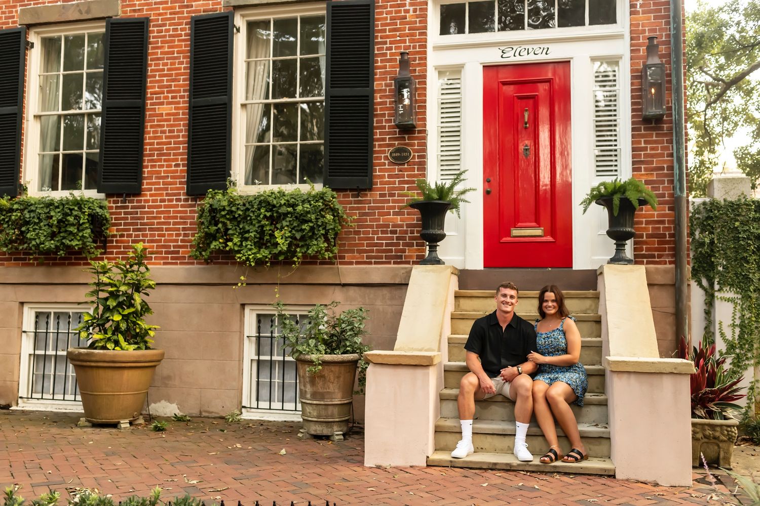 A couple sits on the front steps of a historic red brick townhouse with a bright red door and black shutters in Charleston.