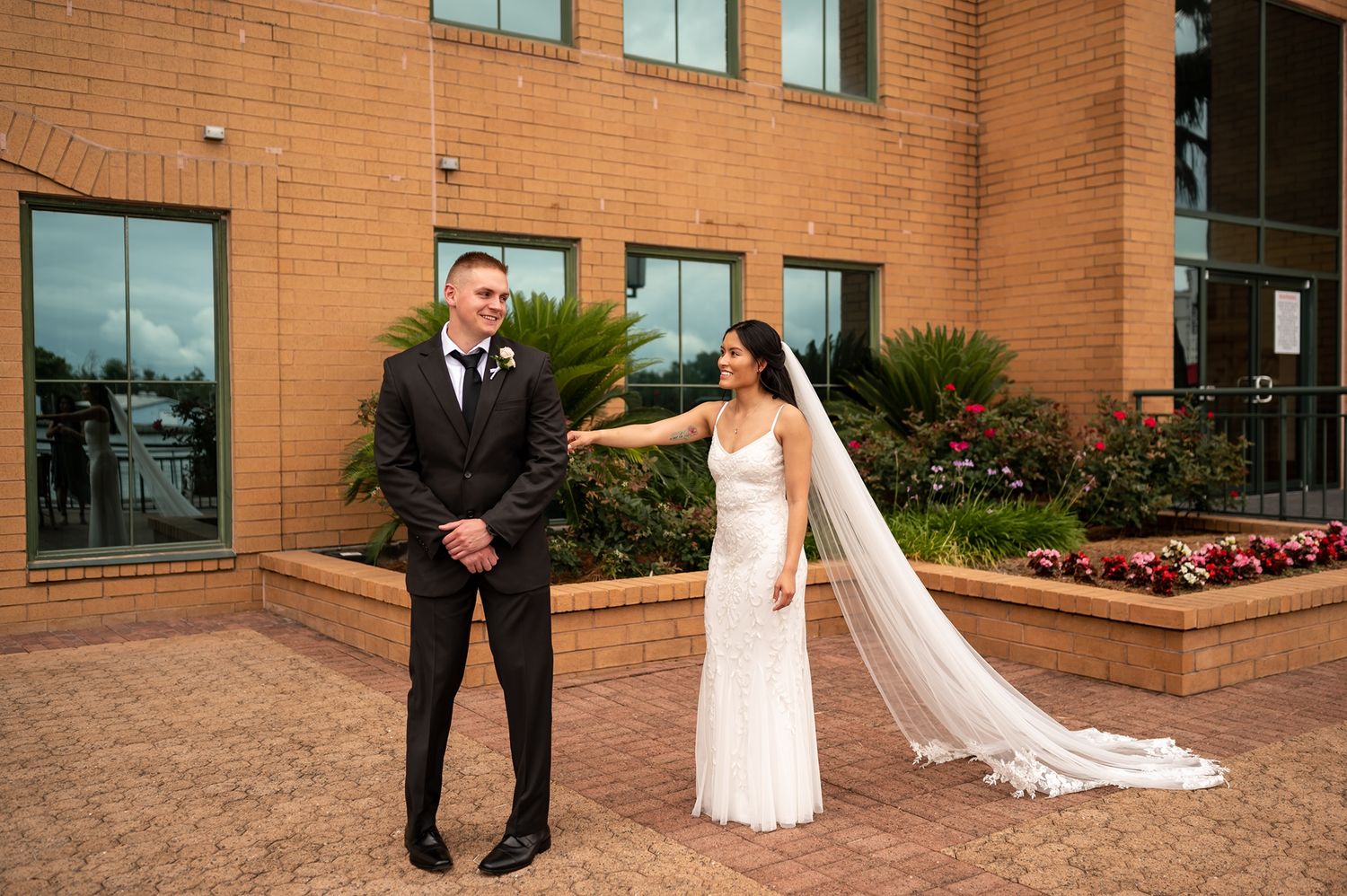 Two people in wedding attire share a happy moment together outdoors in front of a brick building.