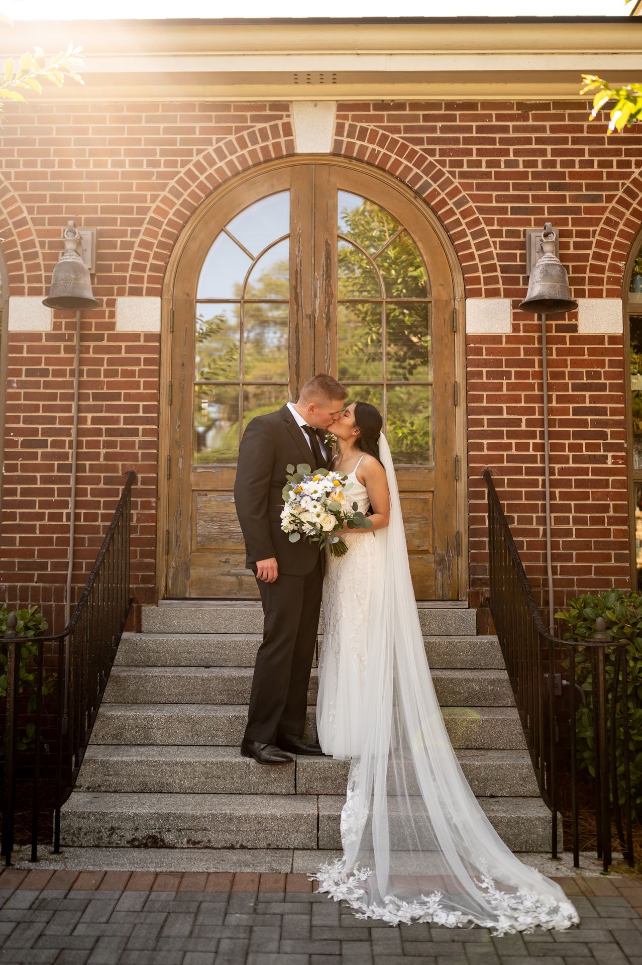 A couple shares a romantic wedding kiss on stone steps in front of an arched brick doorway with vintage lanterns.