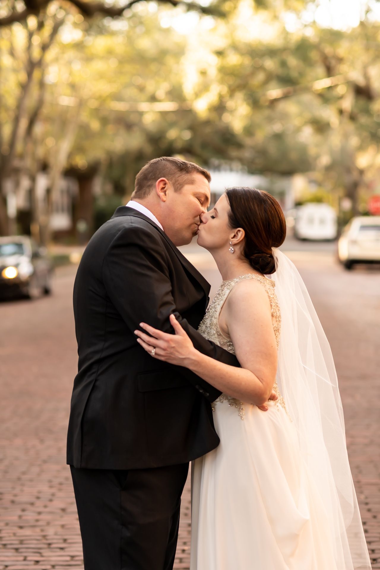 Newlyweds share a romantic kiss on a brick-paved street during golden hour with a flowing white veil and formal attire.