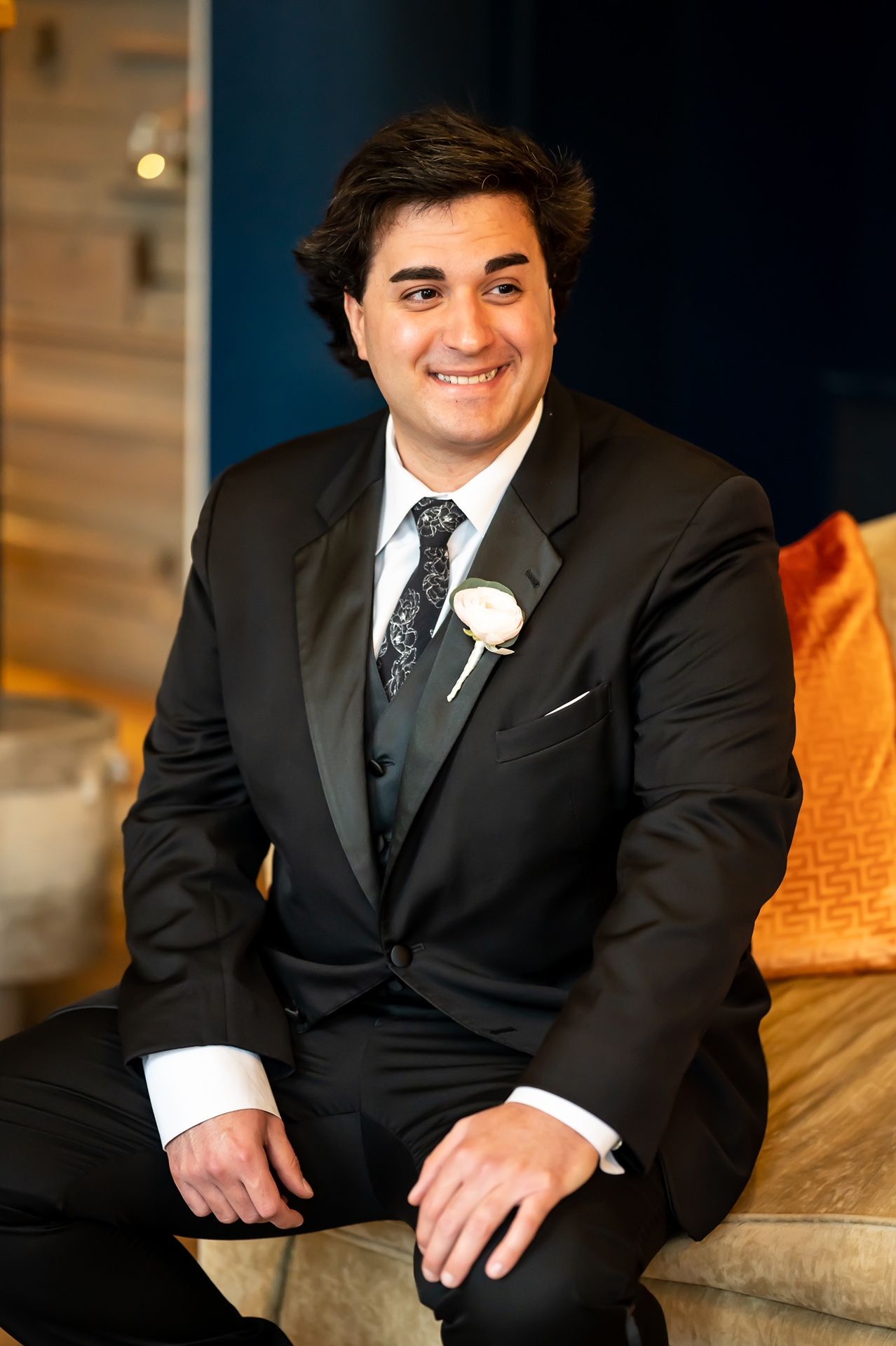 Groom in classic black suit and tie sits on tan couch during formal wedding photography session.