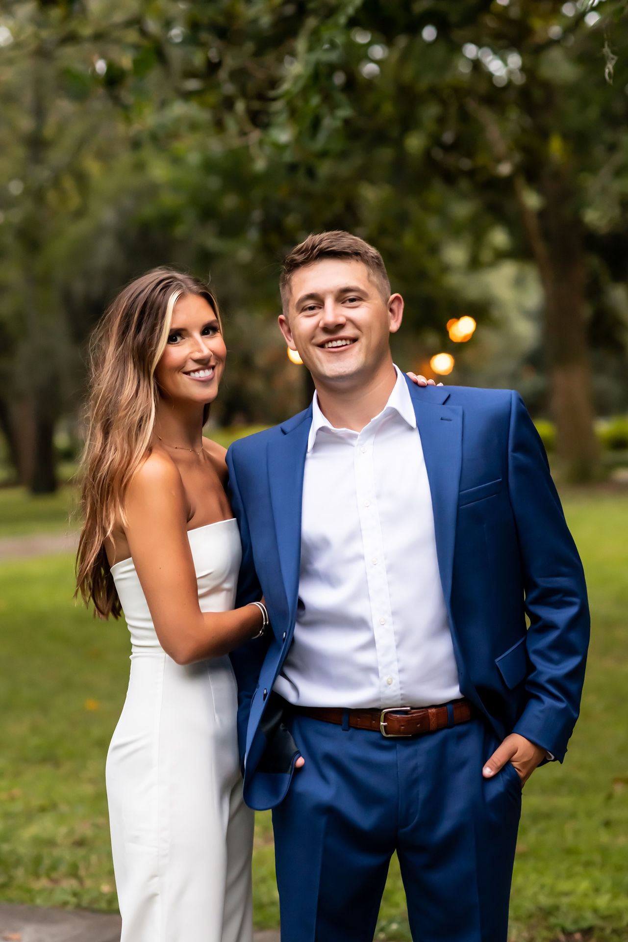 A couple in formal attire smiles together outdoors with a blurred green background and soft lighting.
