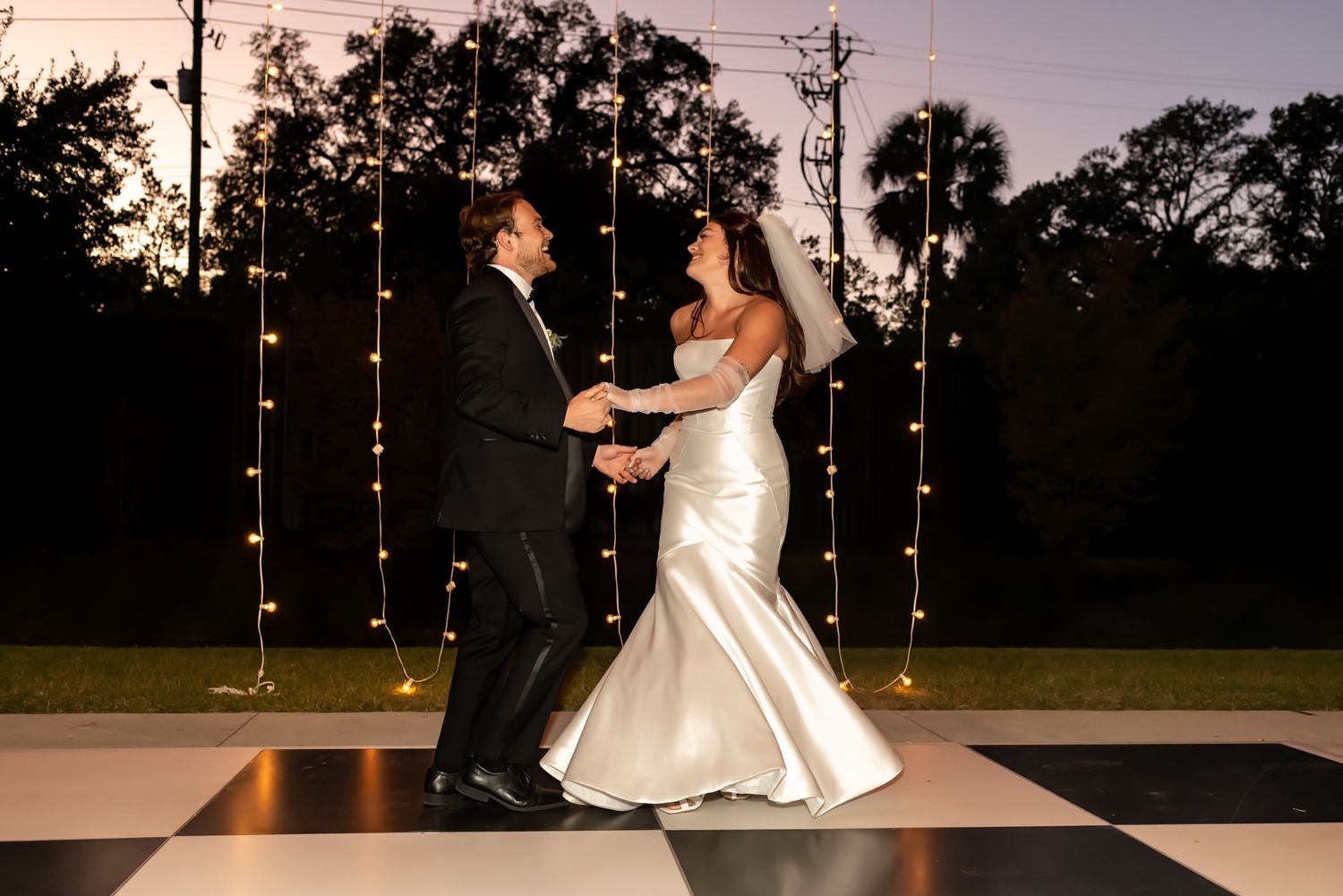 Couple shares romantic first dance together on outdoor dance floor illuminated by string lights at dusk.