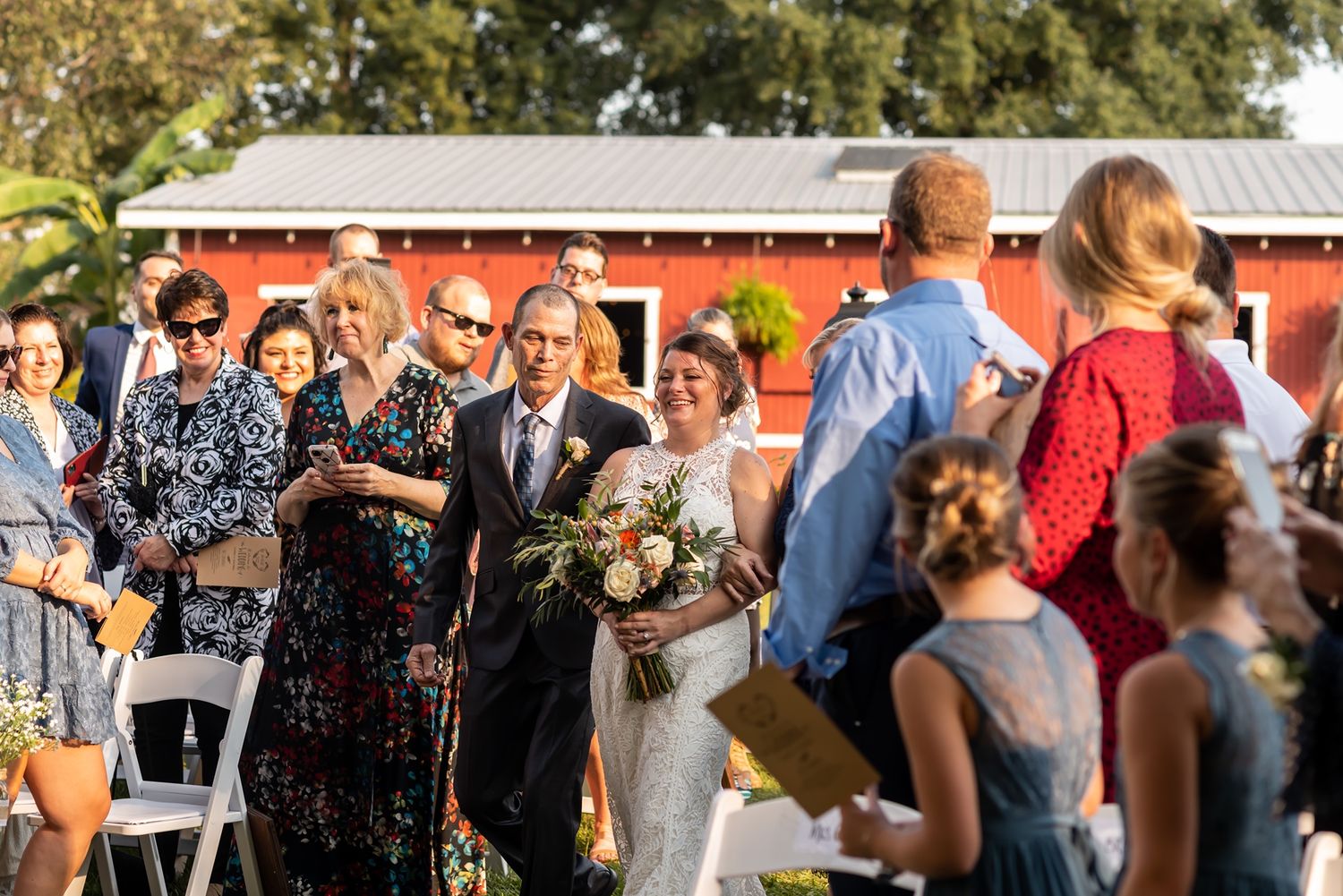 Guests gather outdoors at a rustic farm wedding reception with red barn buildings in the background during sunset.