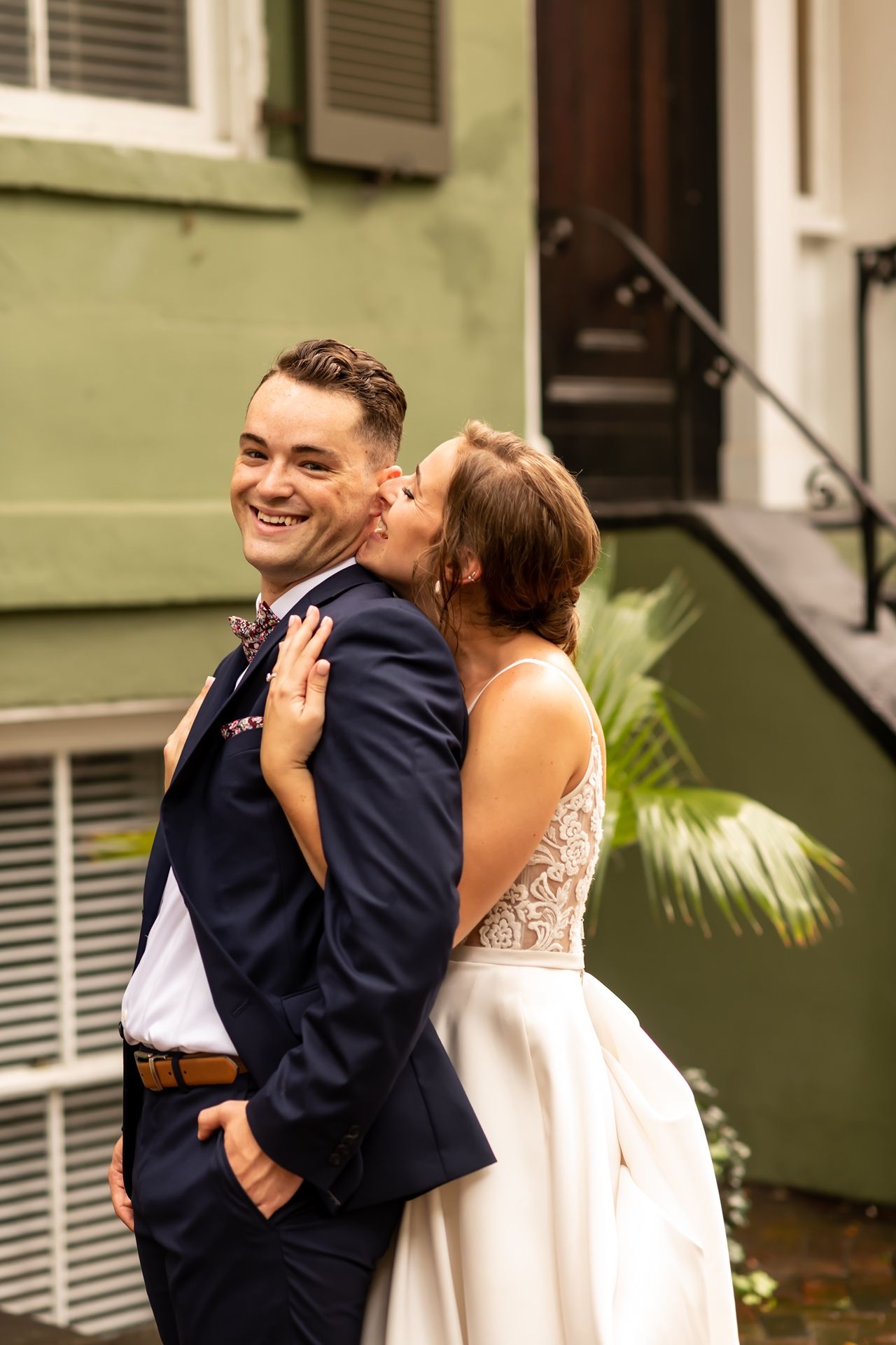 A couple in formalwear embrace tenderly in front of a green building wall during what appears to be a special event.