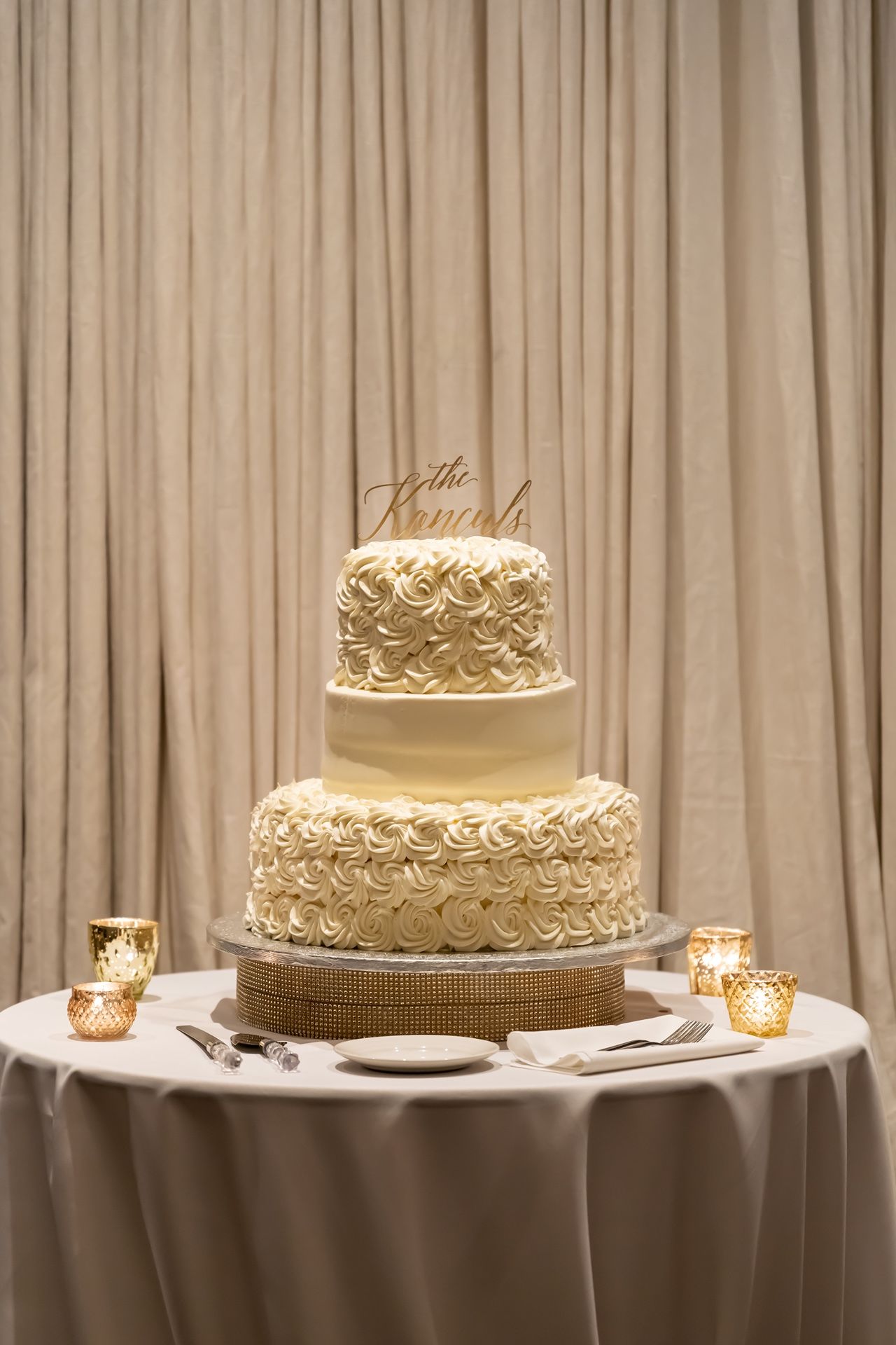 Elegant three-tier white wedding cake with rose swirl decorations on a round table with candlelit votives.