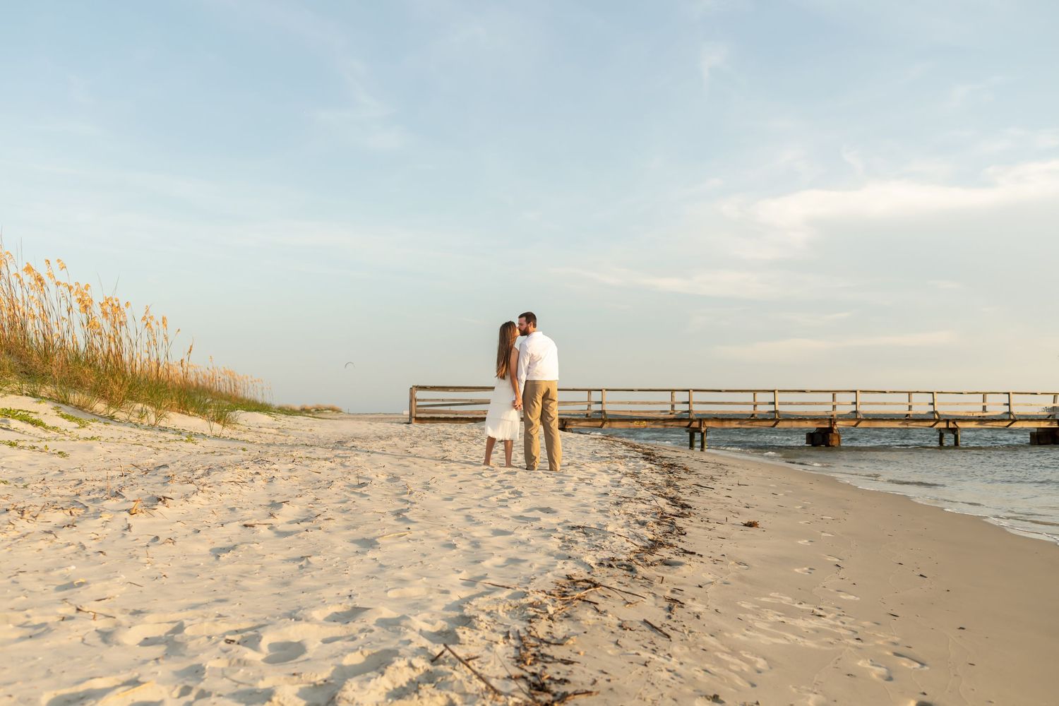 Couple stands together on sandy beach at sunset with wooden pier and sea oats in warm evening light.