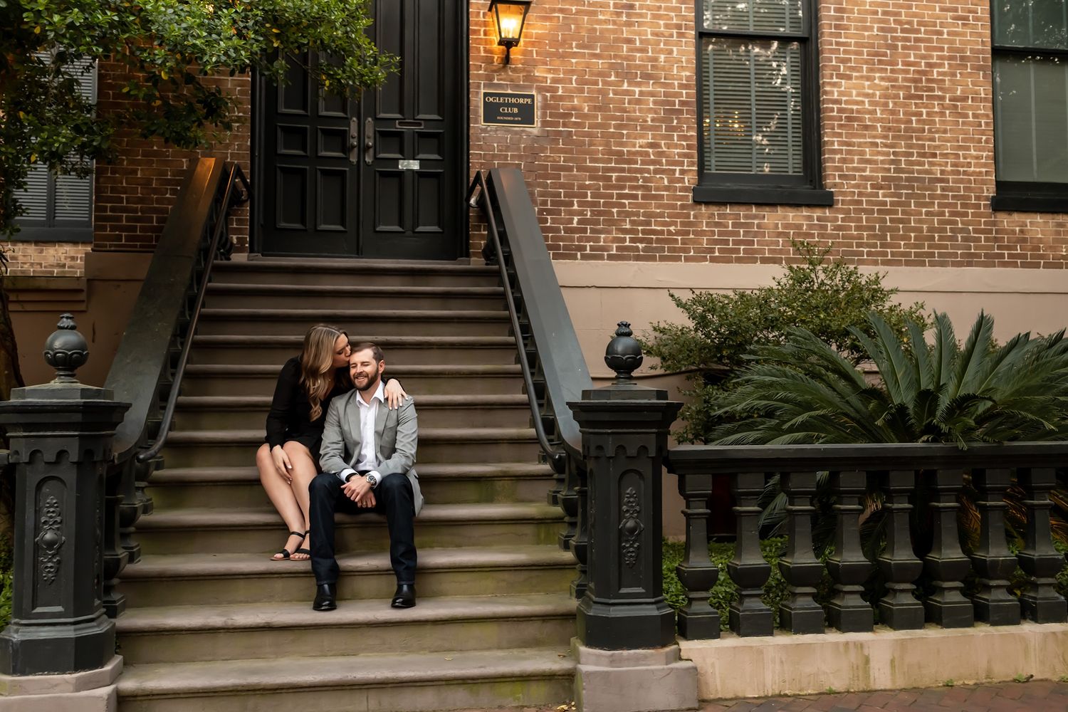 People sit and relax on brownstone steps along a brick building with ornate lampposts and green plants during daytime.