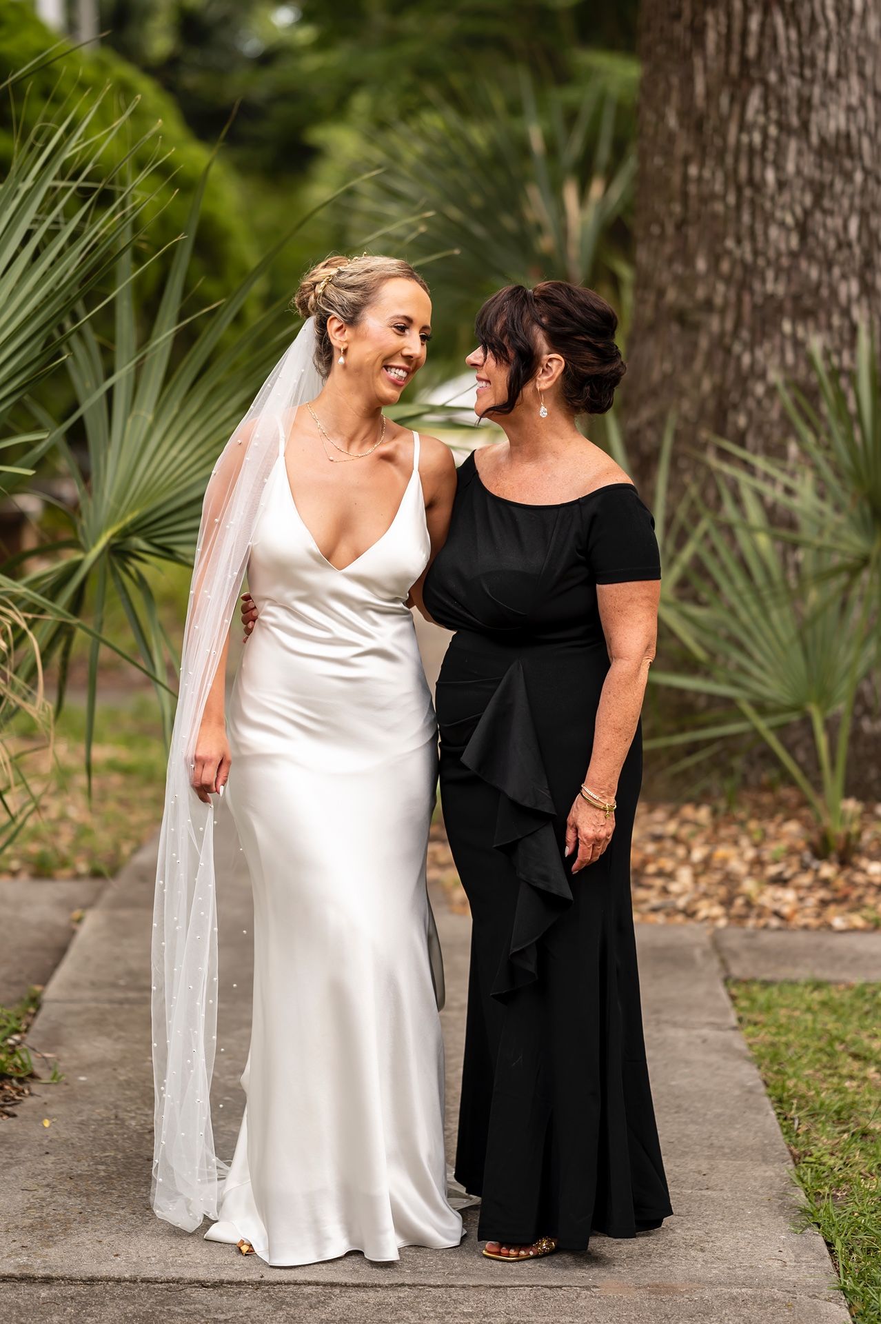 Two people in formalwear share a joyful moment walking together outdoors, one in a white gown and veil, one in black.