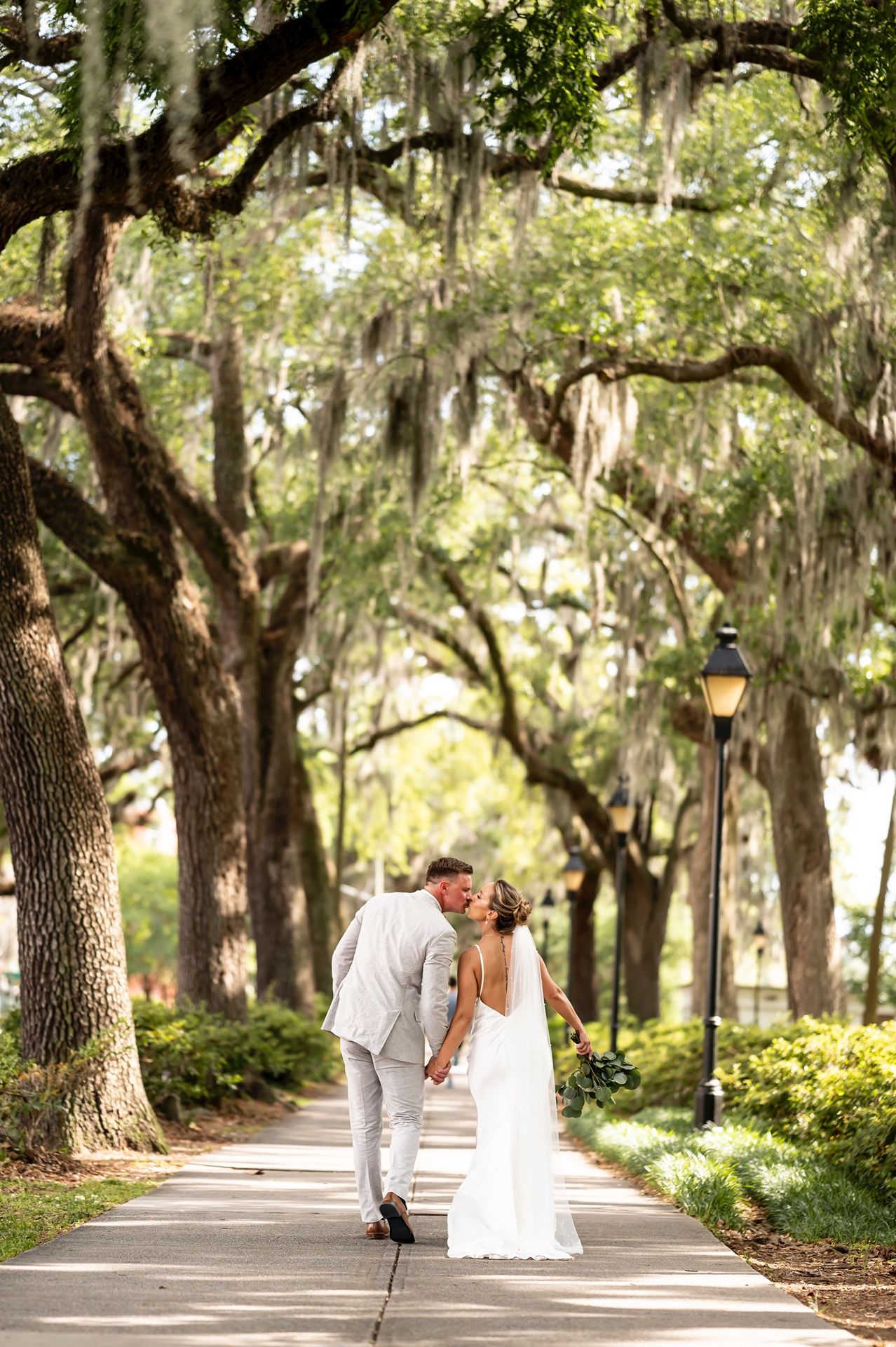 Romantic couple walks along a scenic tree-lined path with Spanish moss hanging from oak branches and a vintage lamppost.