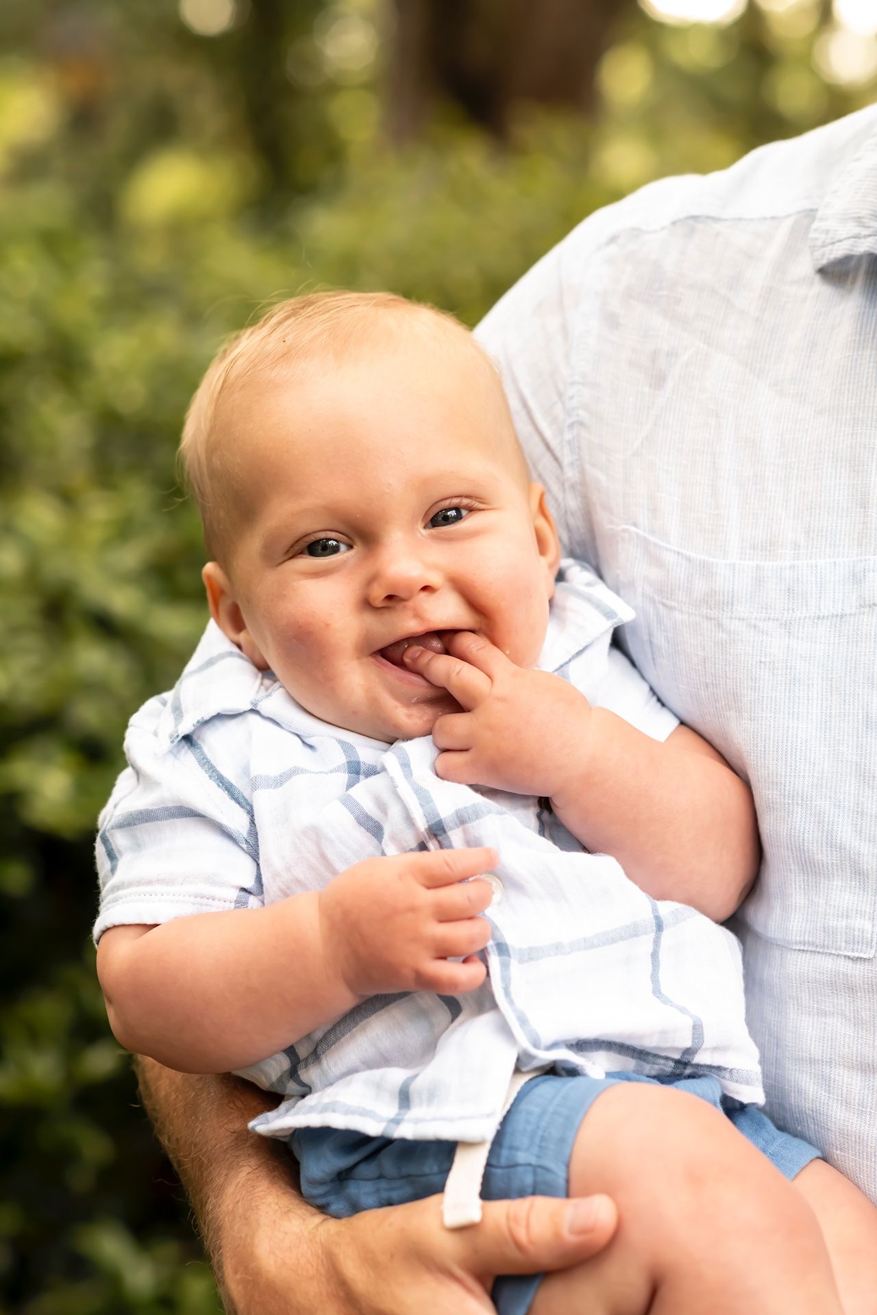 Baby in light blue outfit smiling while being held against a soft blurred background.